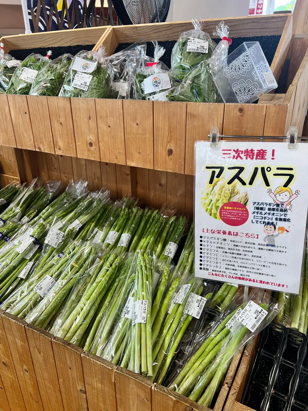 木製の陳列ケースに並んだ、緑色のパプリカと、様々な種類のキノコが置かれている。右側には「アスパラ」と書かれた案内板があり、商品の特徴が説明されている。
