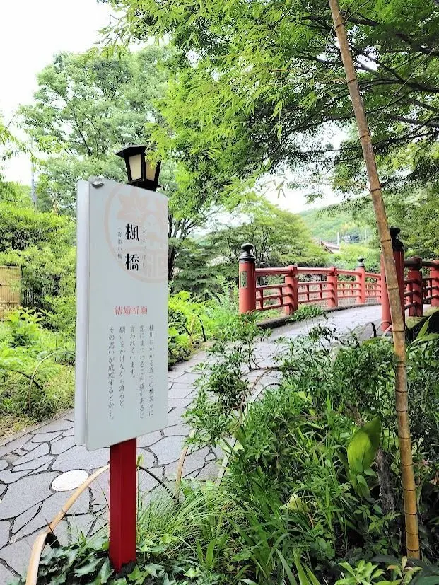 竹や木々が生い茂る庭園の小道、赤い欄干の橋、案内看板が立つ緑豊かな風景
