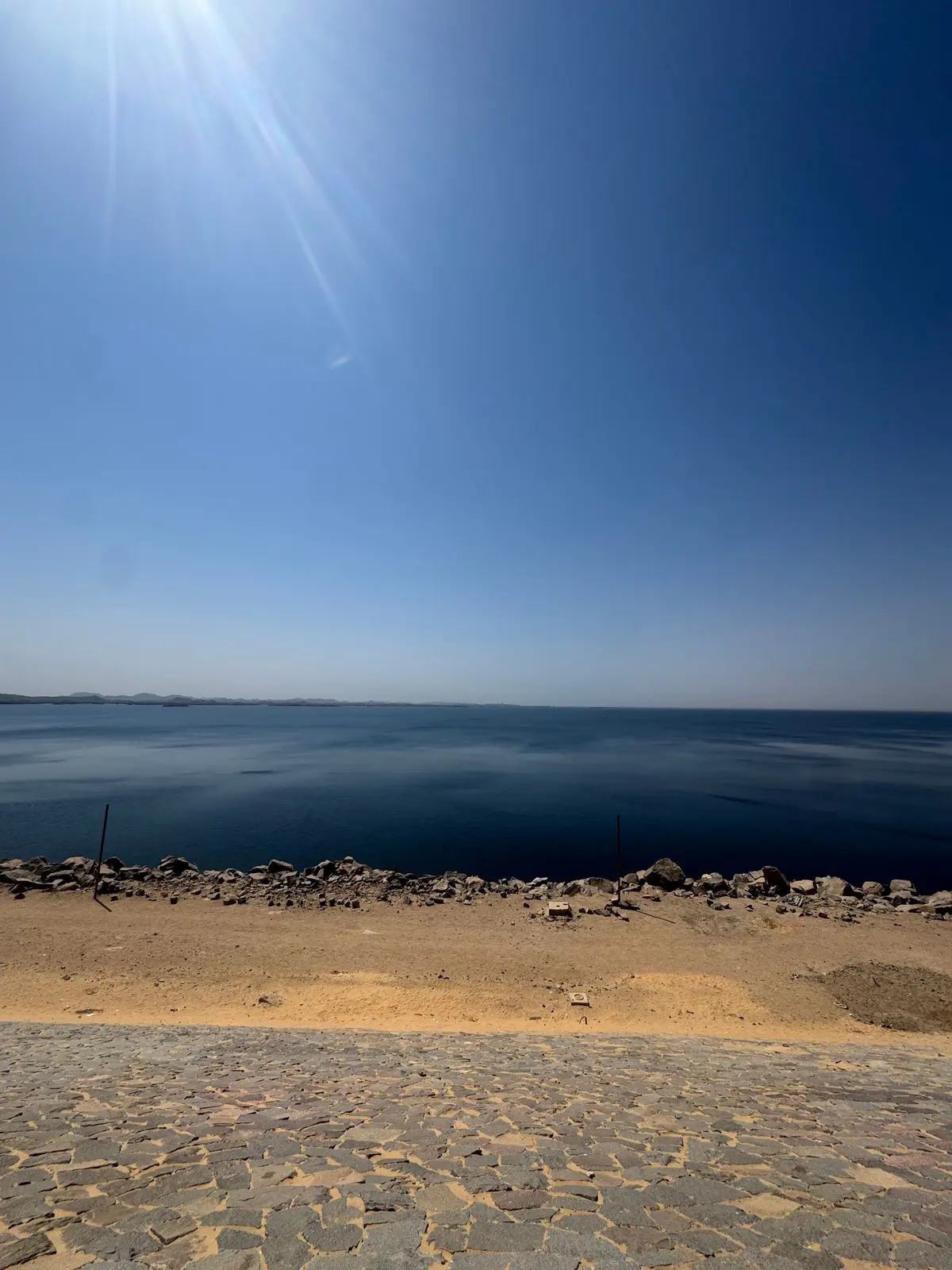水平線が広がる晴天の海辺。手前は石畳の道、その先に砂浜と岩場が続き、遠くにはかすかに陸地が見える。太陽光が強く、空は鮮やかな青。