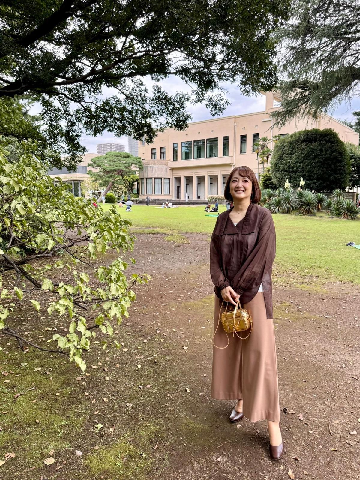 公園の緑道に立つ女性。茶色のブラウスにベージュのスカート、パンプスを着用。右手に小さなゴールドのバッグを持つ。背景には芝生広場とクリーム色の建物が見える。