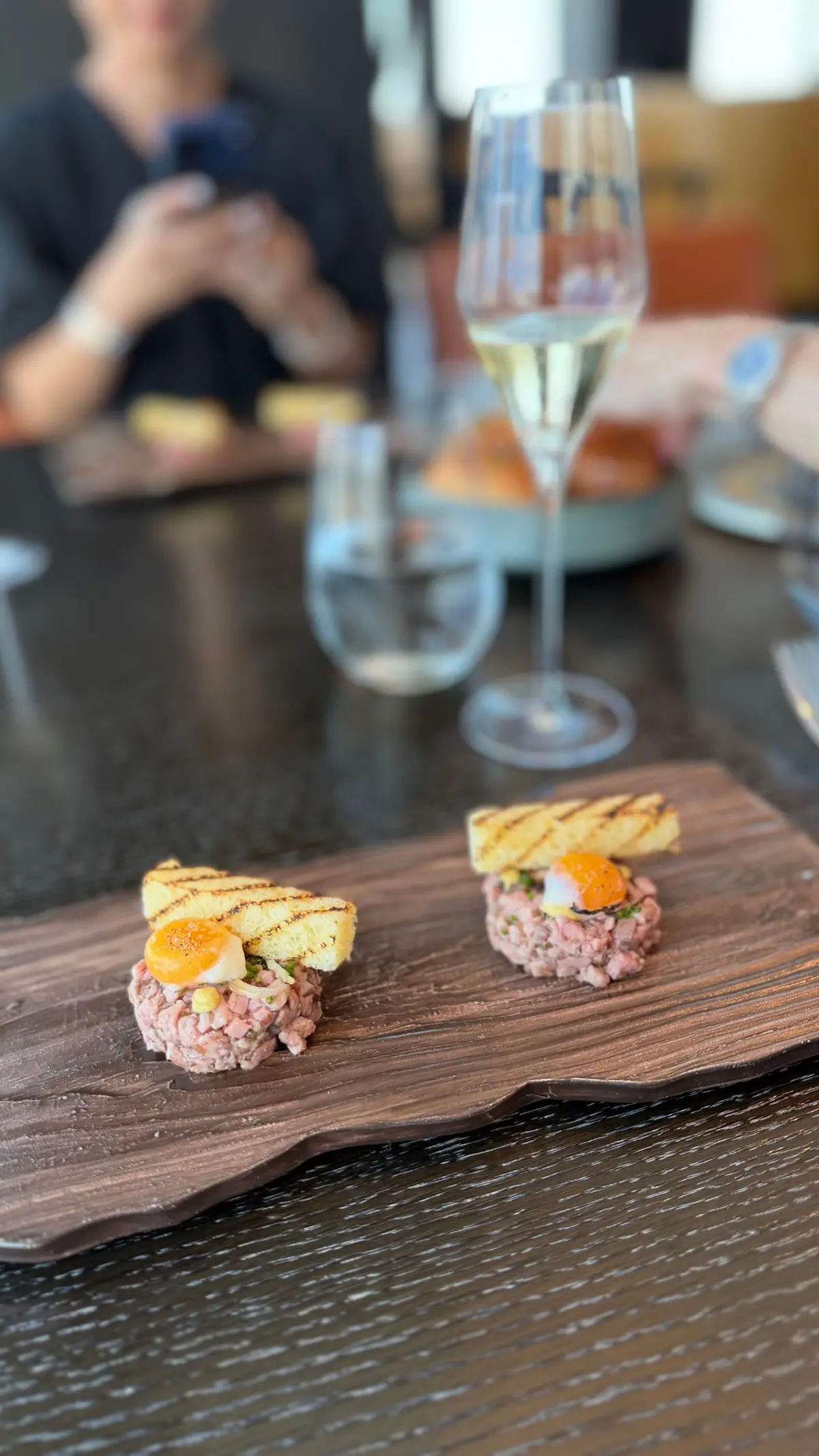 木製の皿に盛り付けられた、牛肉のタルタル2つ。それぞれに黄色のソースと、焼いたトーストが添えられている。背景にはワイングラスと人物がぼやけて写っている。