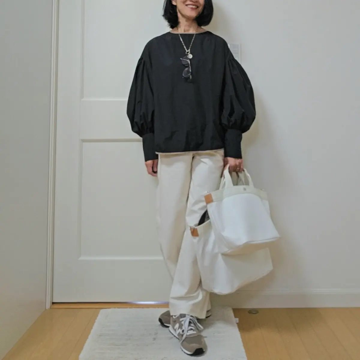 黒いバルーン袖のトップスに白いパンツを合わせた女性。グレーのスニーカーを履き、白いトートバッグを持つ。ネックレスを着用し、笑顔を見せている。背景には白いドアがある。