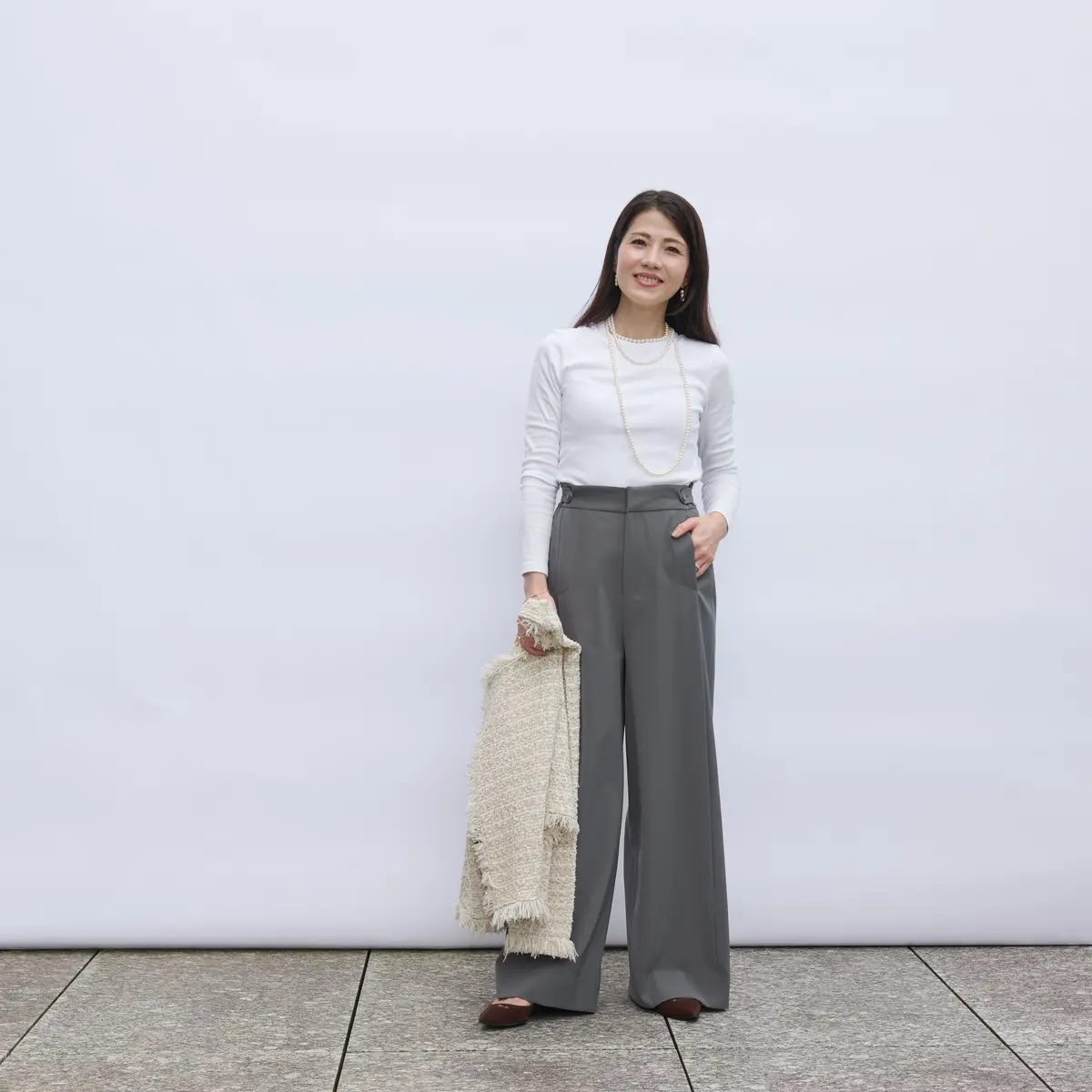 白い長袖シャツにグレーのワイドパンツを着用した女性。右手にベージュのショールを持ち、首には真珠のネックレスをしています。背景は白い壁で、床はグレーの石畳です。