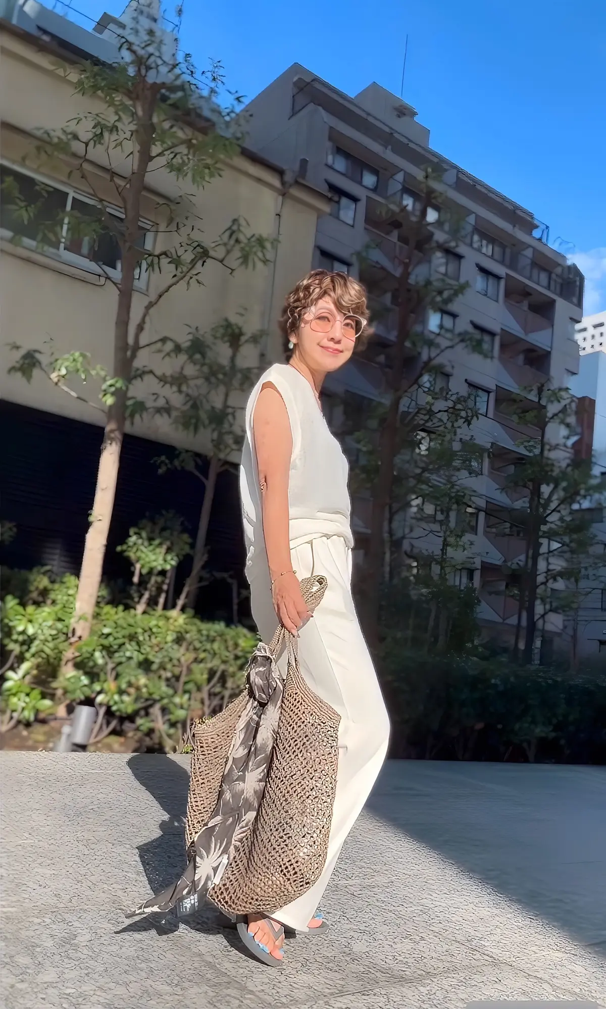 白いタンクトップとパンツを着用した女性が、屋外の歩道で微笑んでいる。茶色の眼鏡をかけ、肩に編み物のバッグをかけている。背景には、ベージュの建物と緑の木々が見える。