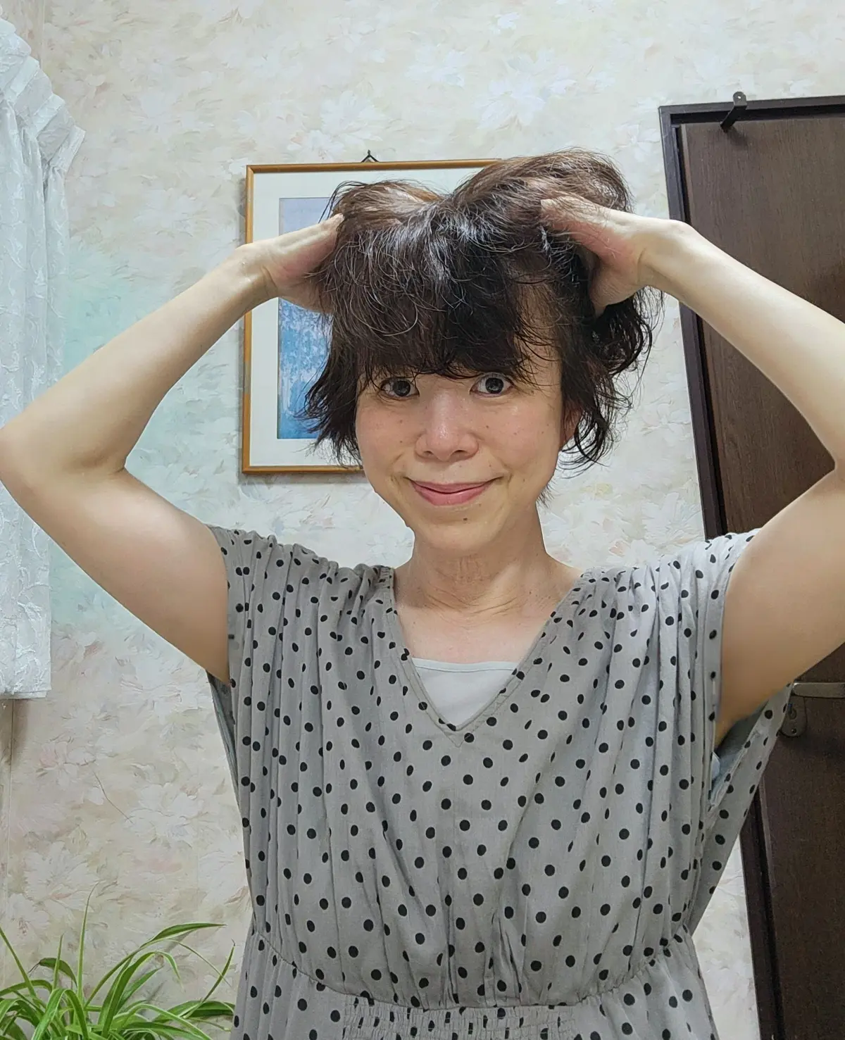 女性が両手を頭に当てて微笑むポートレート。茶色の短髪で、グレーのドット柄ワンピースを着ている。背景には淡い色の壁と絵画、窓帘が見える。