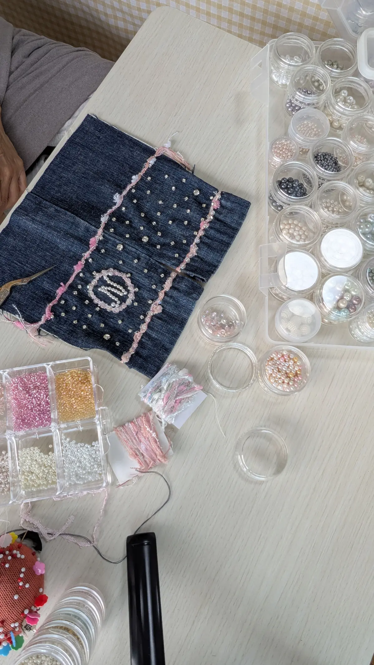 濃い青色のデニム生地に、ピンク色の糸で刺繍が施された作品。中央には白い文字で「S」の文字が刺繍されている。作業台には、大小さまざまな容器に入ったパールやビーズ、刺繍糸が並び、作業道具と思われる黒いペンが置かれている。
