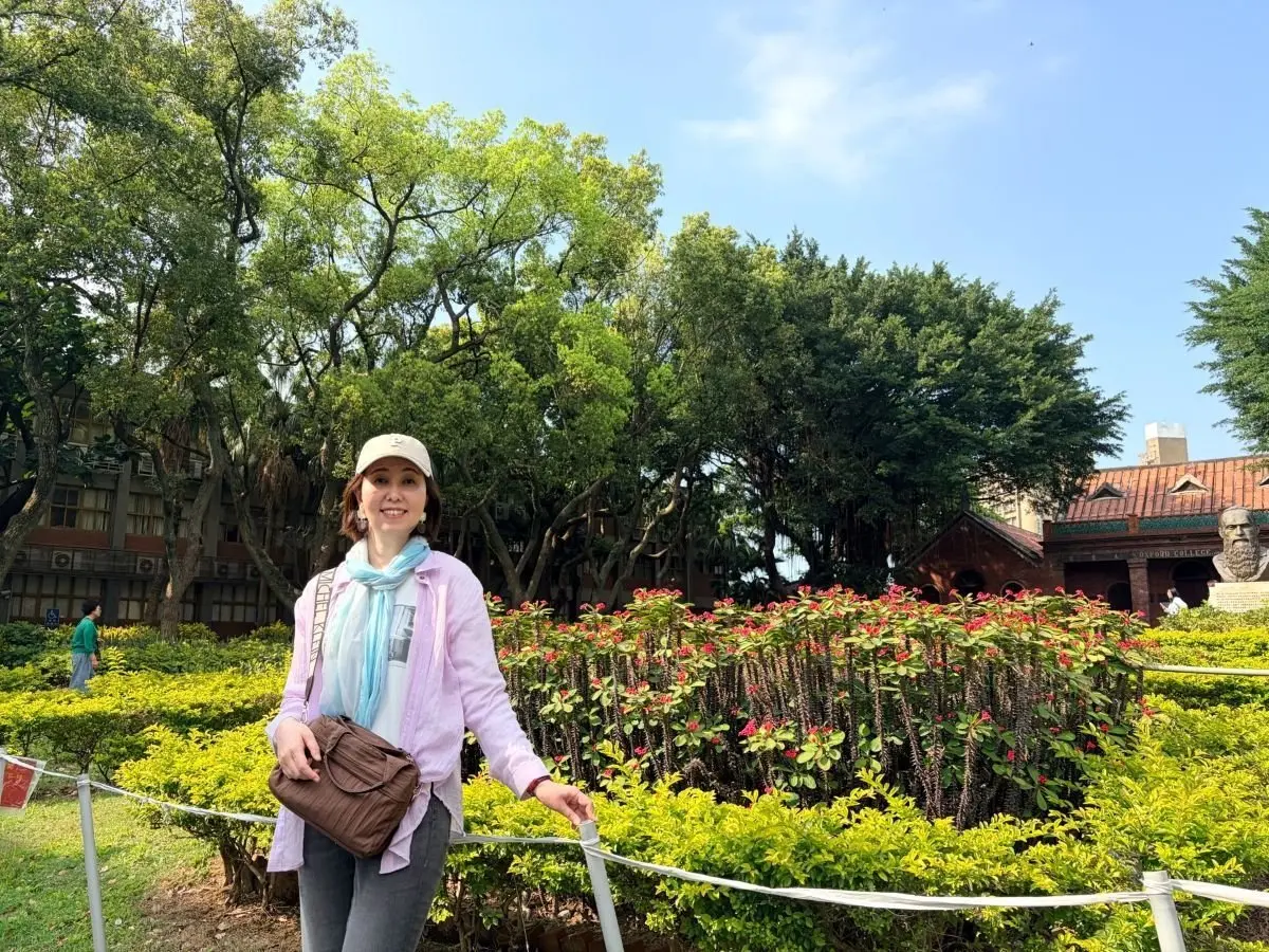女性がピンクのジャケットとキャップで、花壇の前で立ち、笑顔でカメラに向かっています。手には茶色のショルダーバッグを持ち、背景には緑豊かな木々や伝統的な建物が見えます。