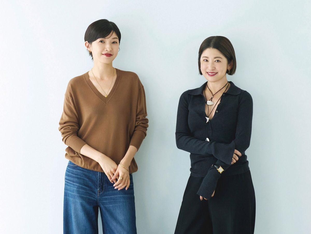 白い壁の前に立つ、茶色のセーターとデニムを着用した女性と、黒いシャツとパンツを着た女性。両者とも短髪で、微笑んでいる。