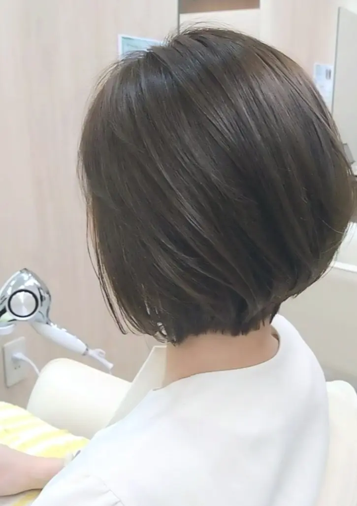 白い椅子に座った女性の後ろ姿で、ショートカットの黒髪が特徴的。白いエプロンを着用し、歯科用の器具が近くに設置されている。