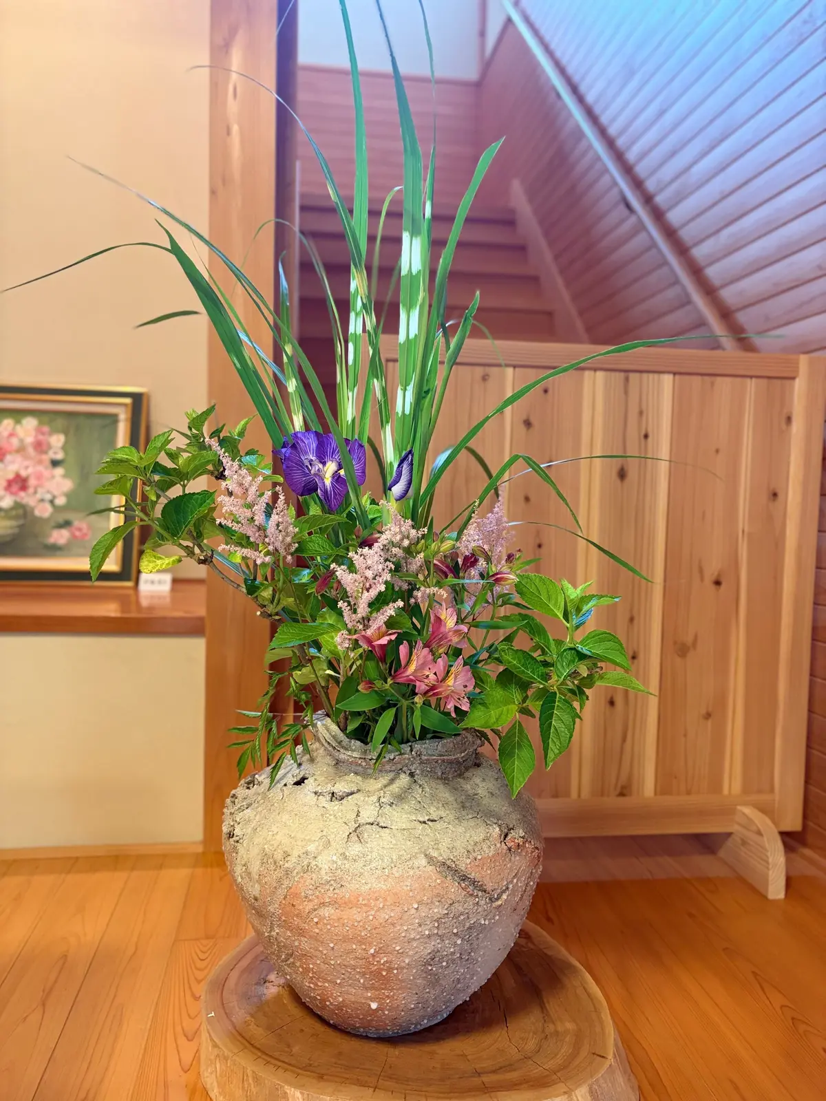 花と緑の植物が並ぶ花瓶、木製の台に置かれた土製の花瓶、背景に絵画と木製の壁