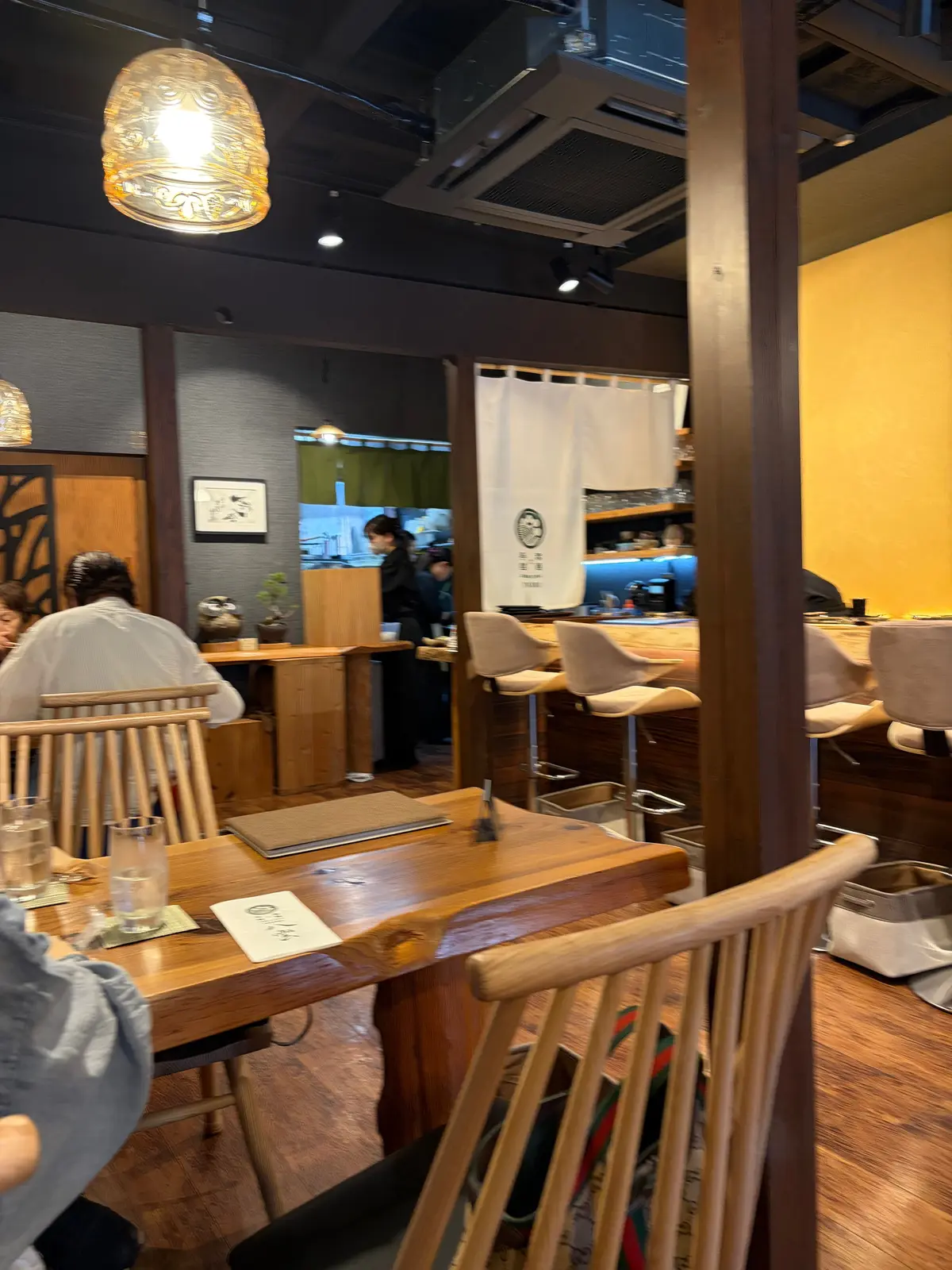 木製のテーブルと椅子が並ぶレストランの内装で、カウンター席とテーブル席があり、客人が座っている。暖かい照明と木製の家具が特徴で、背景には調理場があり、スタッフが作業している。
