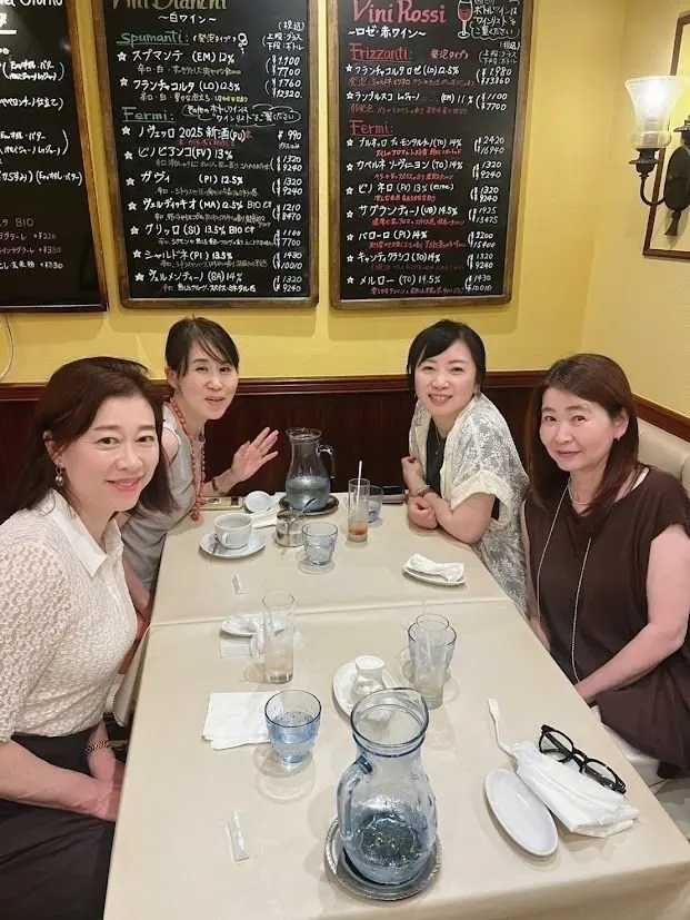 テーブルを囲んで談笑する4人の女性。白いカップに飲み物が注がれ、お茶が入ったポットと水瓶が置かれている。背景にはワインリストが掲げられた壁がある。