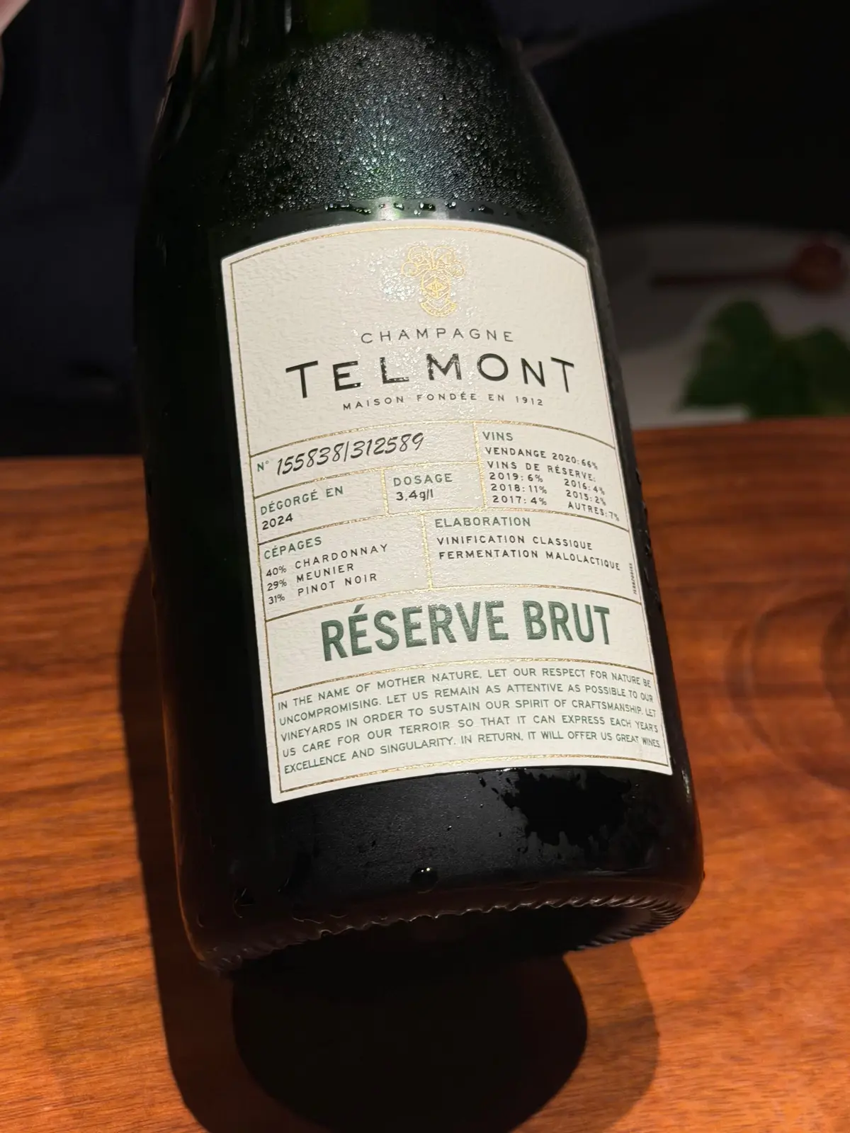 シャンパンのボトルで、ラベルには「TELMONT MAISON FONDÉE EN 1912」とあり、「RÉSERVE BRUT」と記載されている。木製のテーブルに置かれている。