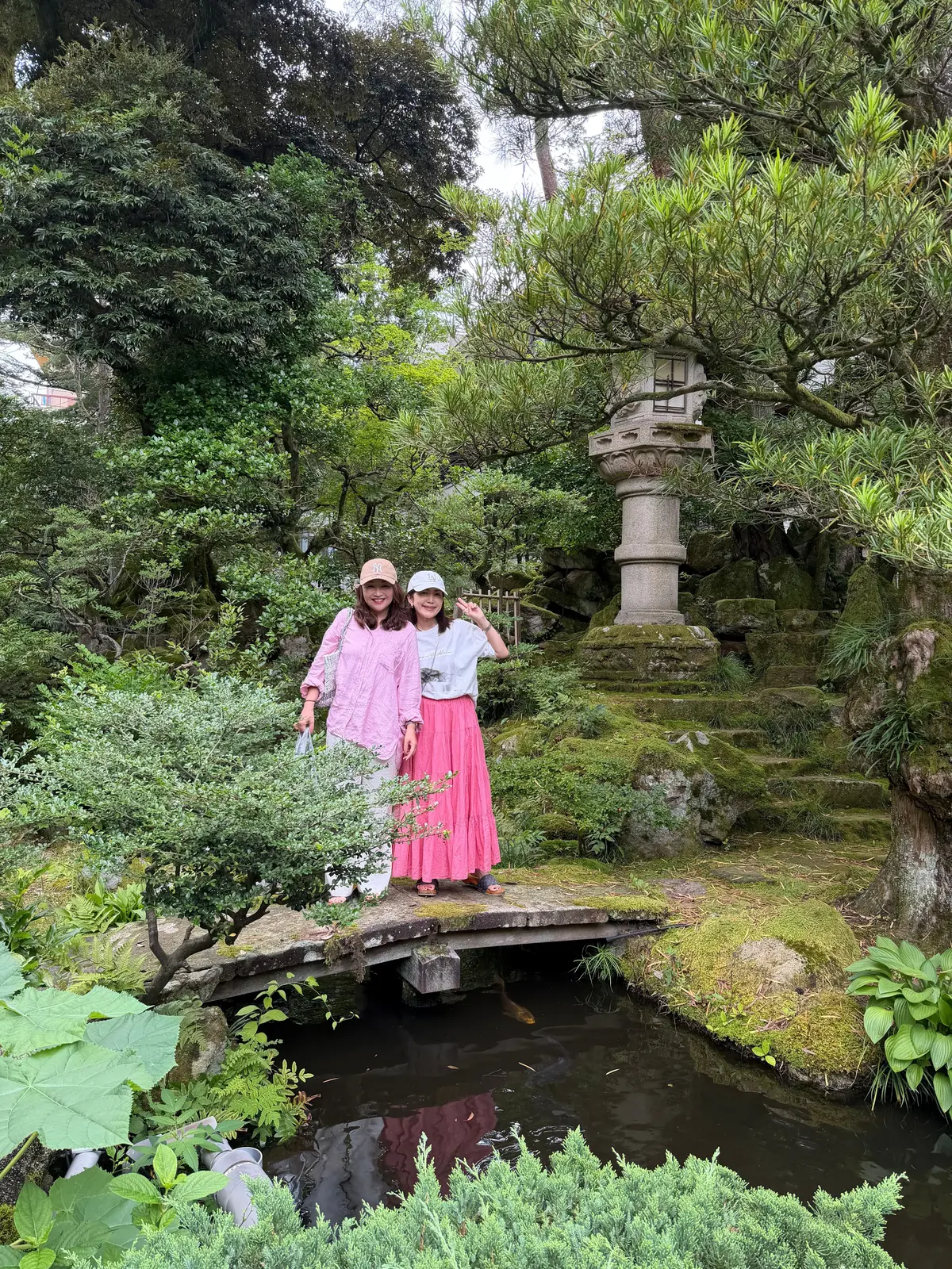 ピンクのトップスとスカートを着た2人の女性が、苔むした石橋の上で笑顔でポーズをとっている。背景には、緑豊かな植栽と石灯籠が見える庭園の風景。