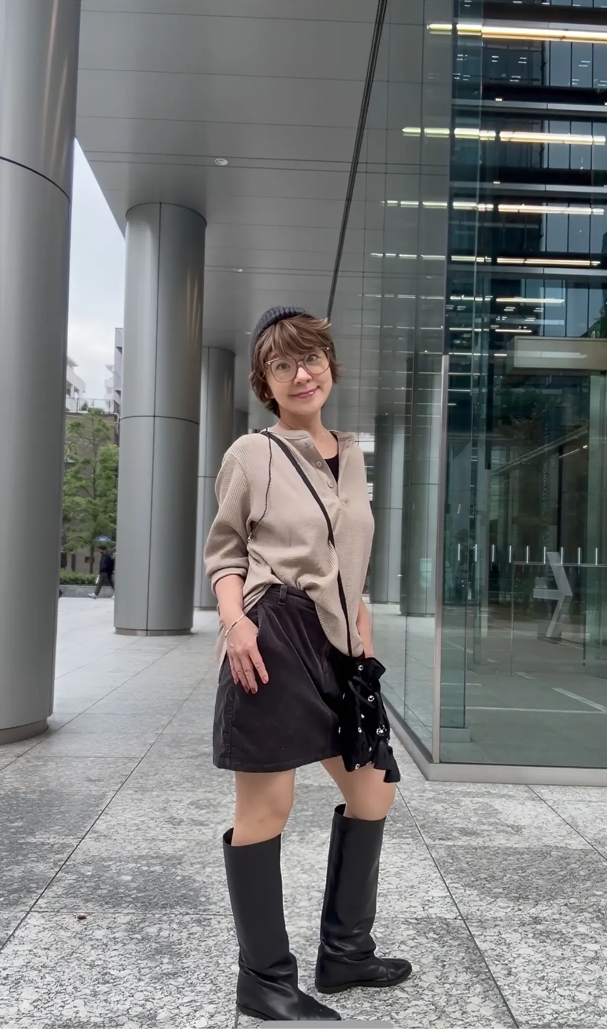 ベージュのカーディガンに黒のミニスカート、ロングブーツを合わせた女性。丸メガネをかけ、笑顔でこちらを見ている。背景は現代的な建築物の外観。
