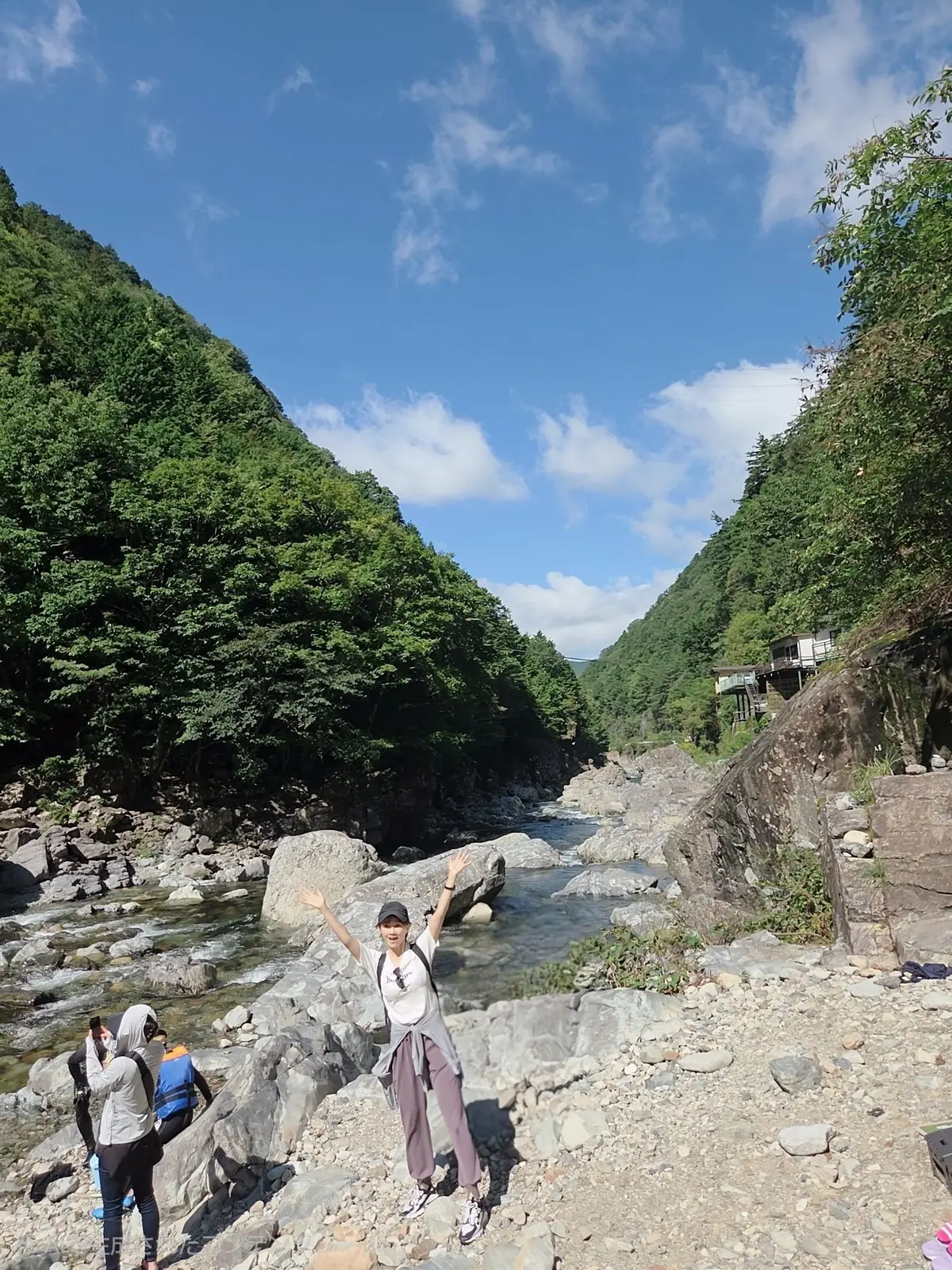 緑豊かな山々に挟まれた渓谷に、浅瀬の川が流れる風景。中央には両手を広げ喜ぶ女性、左には別の女性が立っている。川岸は岩場になっている。