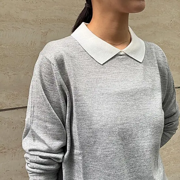 グレーの長袖ニットの上に白い襟が付いたトップスを着用した女性。髪は後ろでポニーテールにまとめられている。背景はベージュ色の壁。