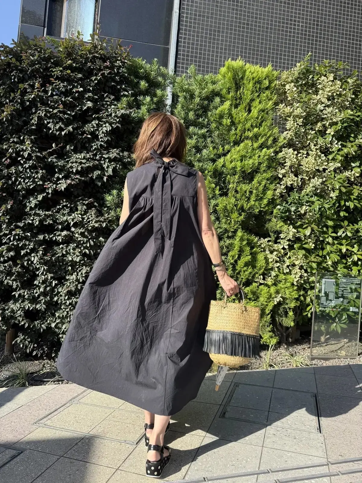 黒のノースリーブワンピースを着た女性が、背面から見え、編みかごバッグを手に持って歩いています。ブッシュの生い茂る背景に、モダンな建物の一部が映っています。