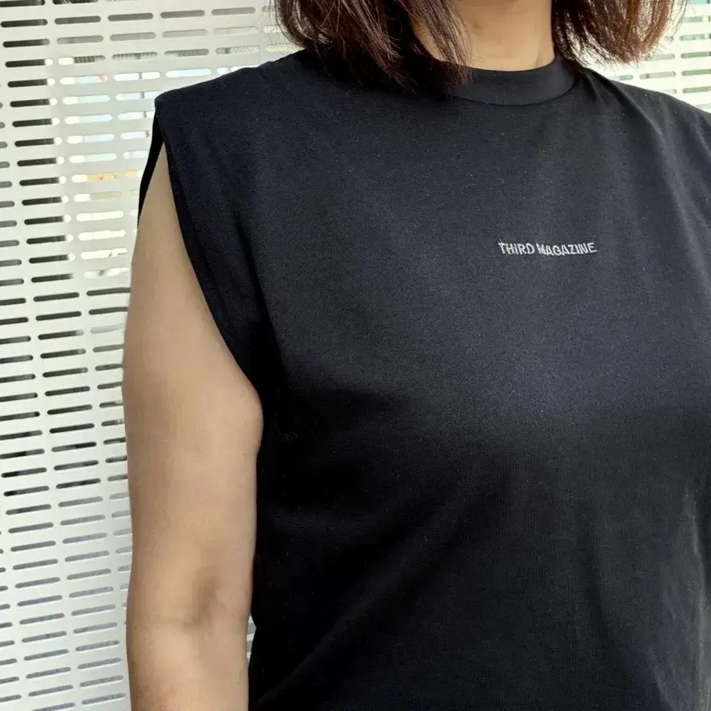 黒いスリーブレスTシャツを着た女性。胸元に白字で「THIRD MAGAZINE」とプリントされている。背景には白いスリットが入った壁がある。
