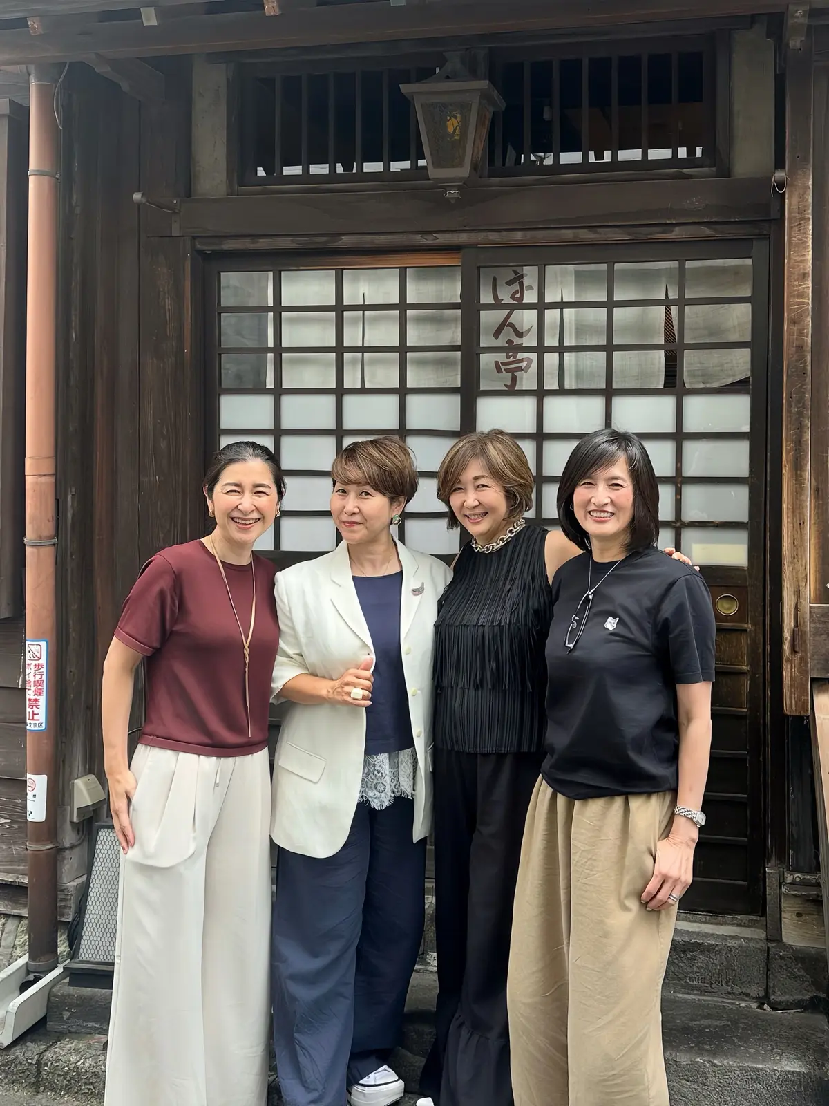 根津、串揚げ「はん亭」、伝統的な木造建築の門前で、4人の女性が並んで立っている。各女性は異なる服装で、左からマロンレッドのトップスと白のスカート、ベージュのコートとネイビーのスカート、黒のトップスと黒のスカート、黒のトップスとベージュのスカートを着ている。