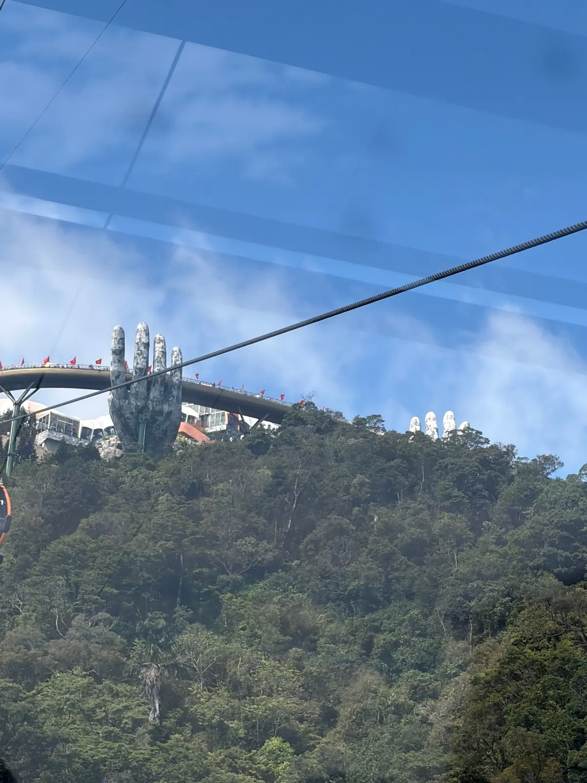 緑豊かな山頂に、巨大な石の手の形をした建造物。橋の上を人が歩き、赤いベトナムの旗がはためいている。青空に白い雲が浮かんでいる。