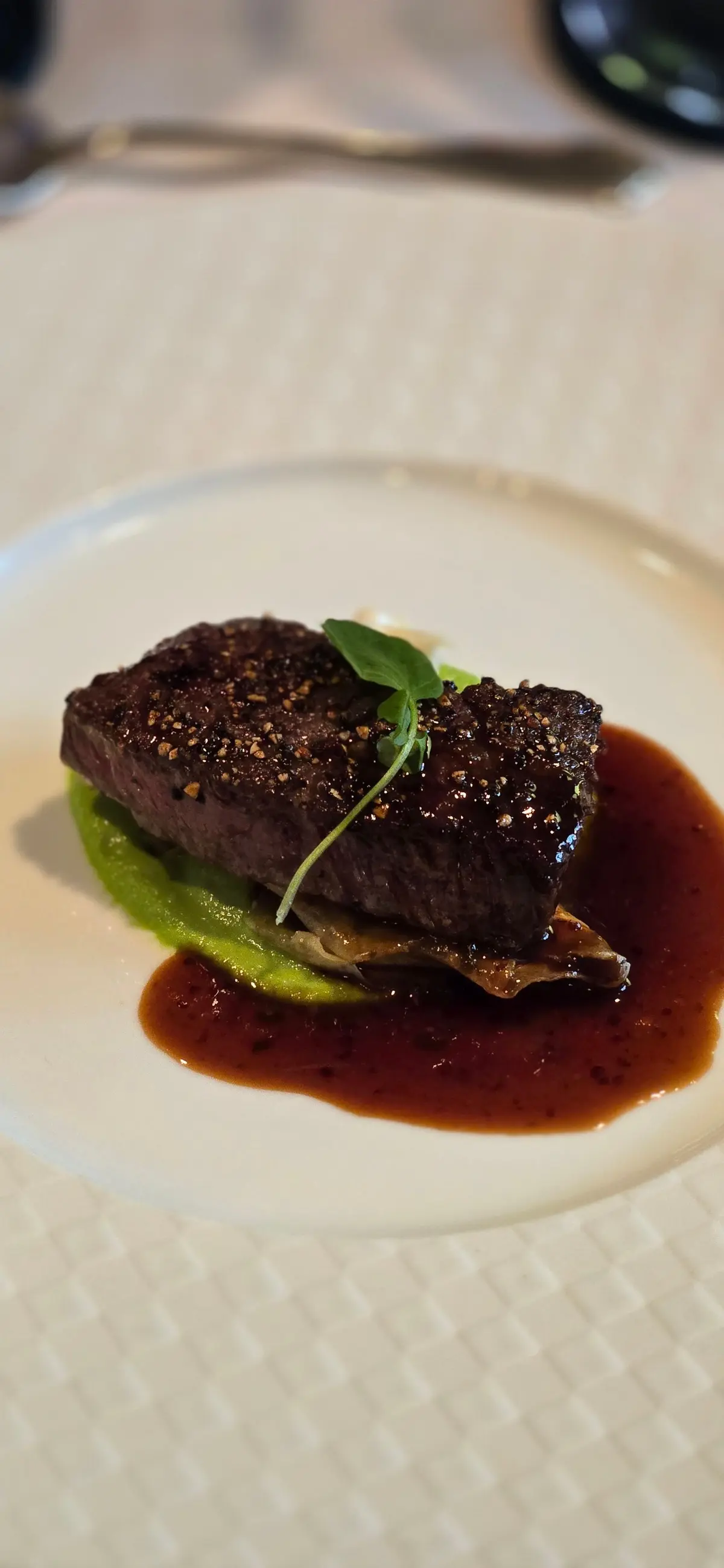 白磁の皿に盛り付けられた、黒胡椒で覆われた牛肉のステーキ。緑色のソースと、濃い赤褐色のソースが添えられ、手前のほうにはキノコが敷かれている。