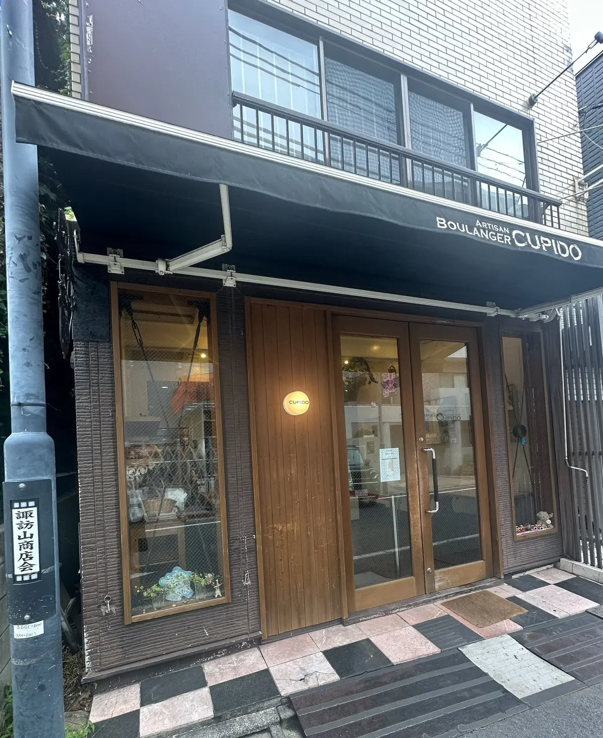 白とグレーの建物に面した、パン屋「カップIDO」の店構え。入り口は木製ドアで、ショーケースにはパンが並んでいる。看板には「ARTISAN BOULANGER CUPIDO」の文字。