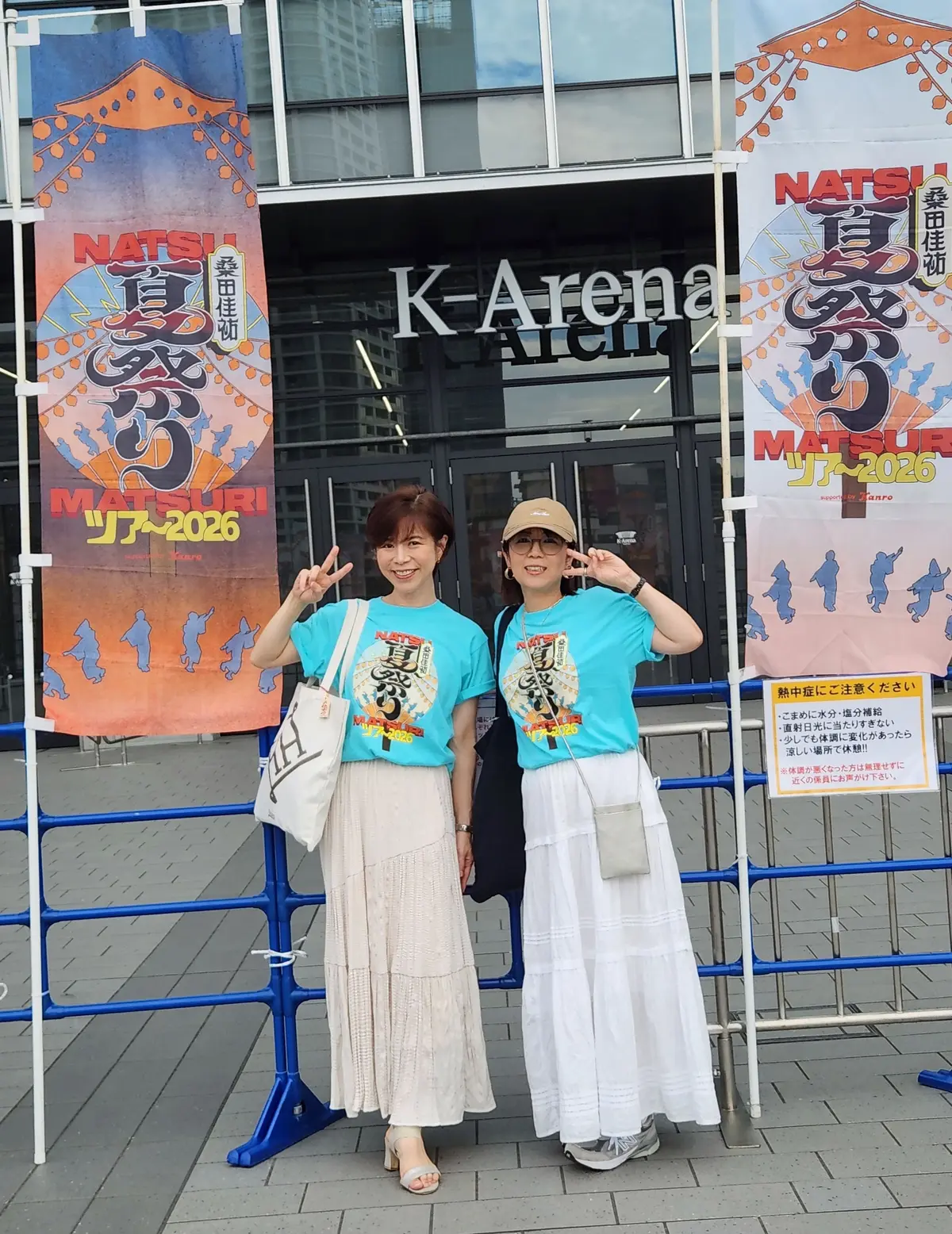 K-Arenaの前で、2人の女性が笑顔で記念撮影をしている。左の女性はオレンジ色のTシャツ、右の女性は緑色のTシャツを着ている。背景には、夏祭りのポスターと提灯が飾られている。