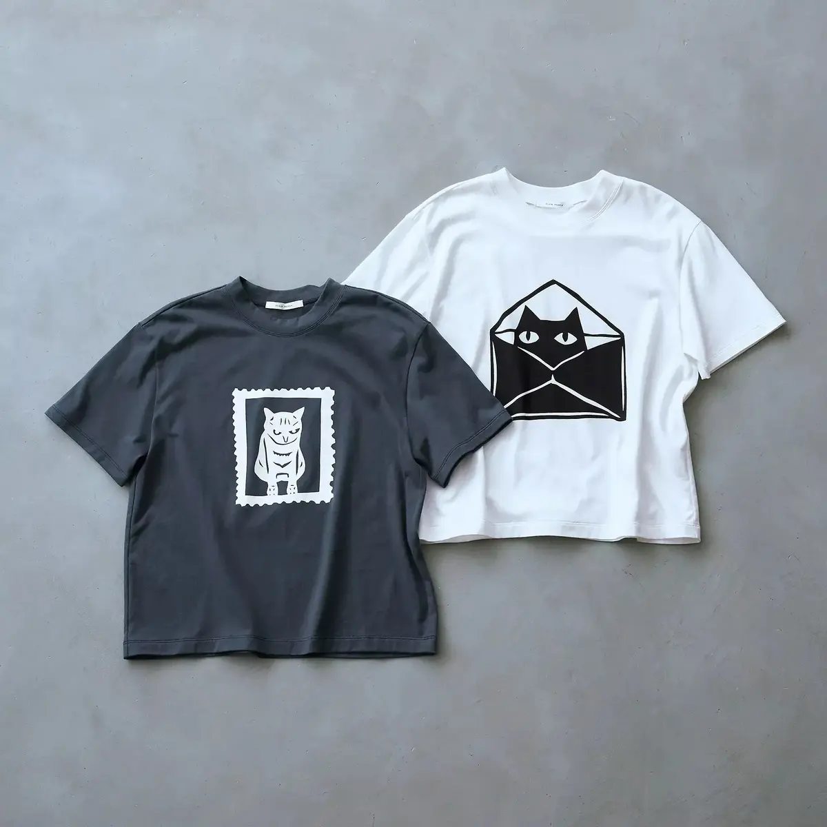 左は濃いグレーのTシャツに白抜きの猫の郵便切手プリント、右は白のTシャツに黒猫が封筒に入ったイラスト。両方ともゆったりとしたデザイン。
