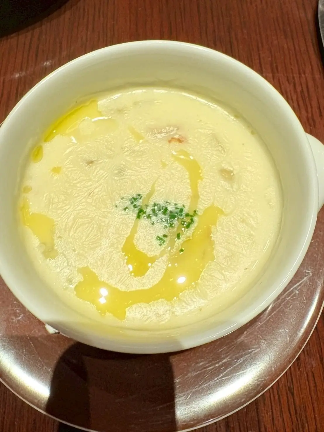 白い器に入った、クリーム色のムースまたはポタージュ。表面にはオリーブオイルと刻みネギが散らされ、木製のテーブルに置かれている。