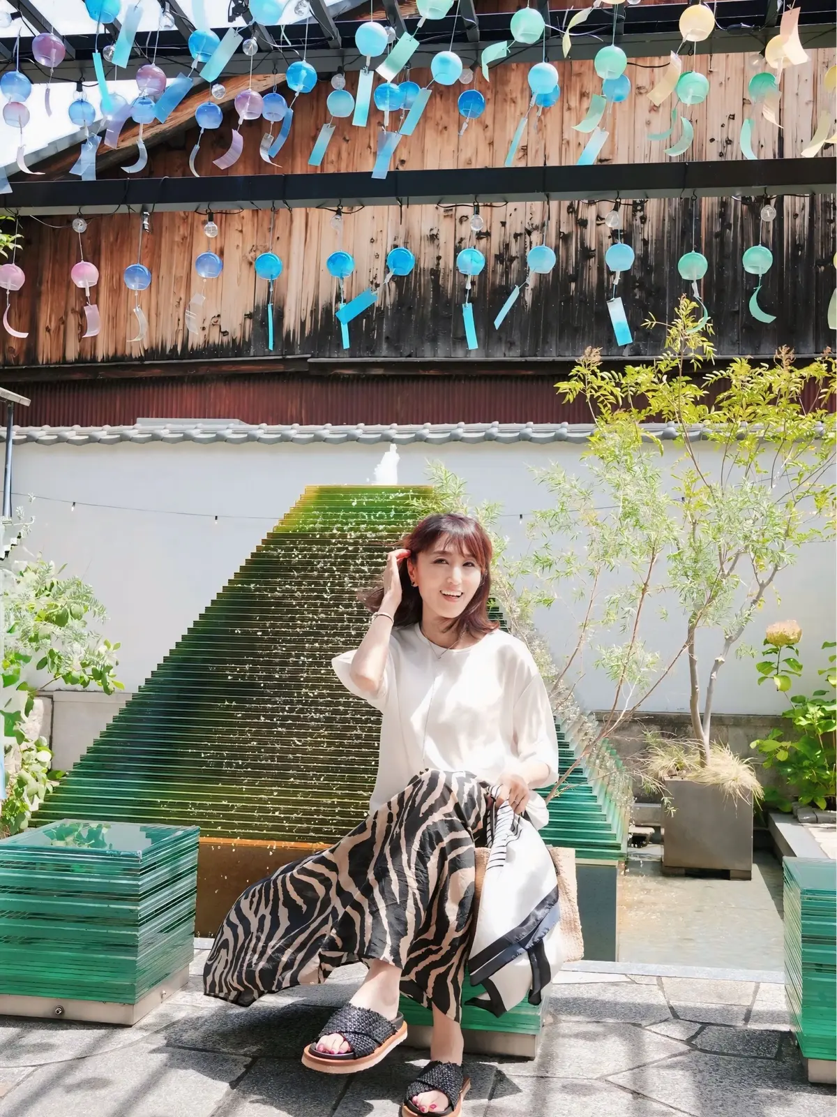 木陰で、白シャツにゼブラ柄のワイドパンツ、サンダルを履いた女性が座っている。背景には青色の階段と、木漏れ日が差し込む天井に飾られた風鈴が見える。