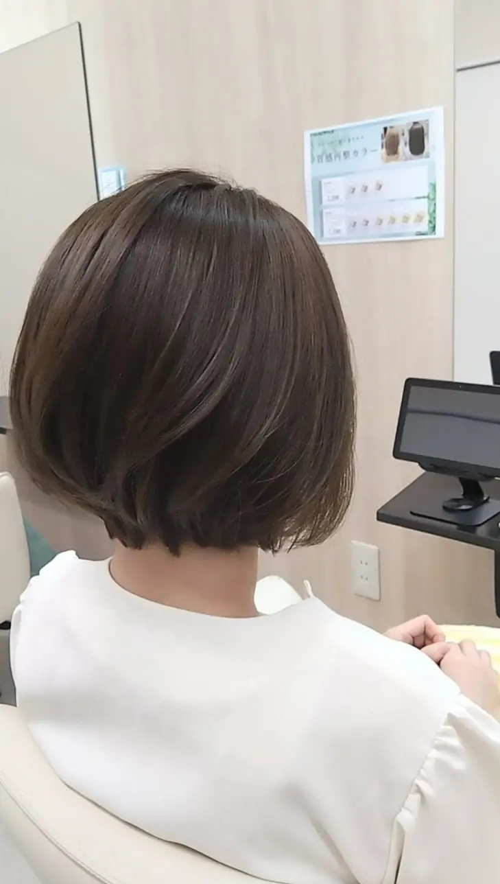 白い椅子に座った女性の後ろ姿で、短い黒髪が特徴的。背景にはメニュー表とモニターが置かれたカウンターがあり、コーヒーカップも見える。