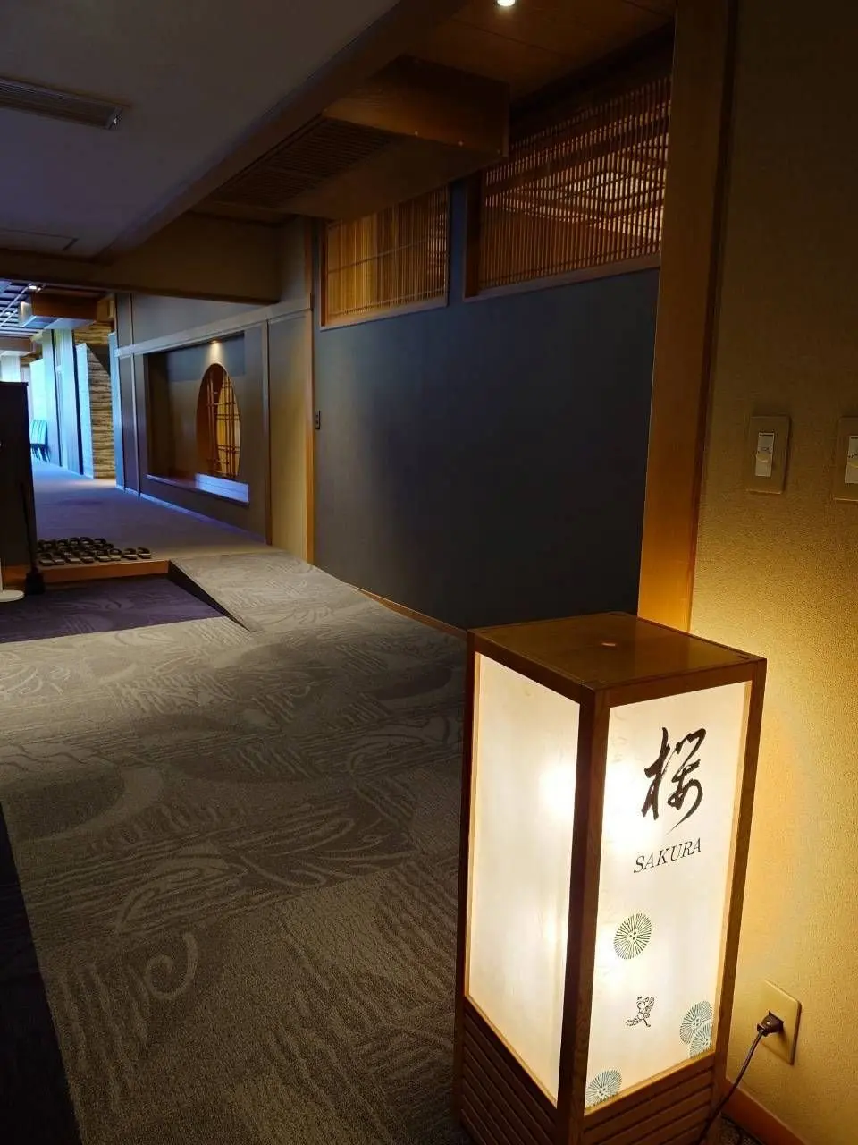 常磐ホテルの廊下。右には「SAKURA」と書かれた照明が置かれ、床には波模様の絨毯が敷かれている。背景には障子や木製の装飾が施された通路が続く。