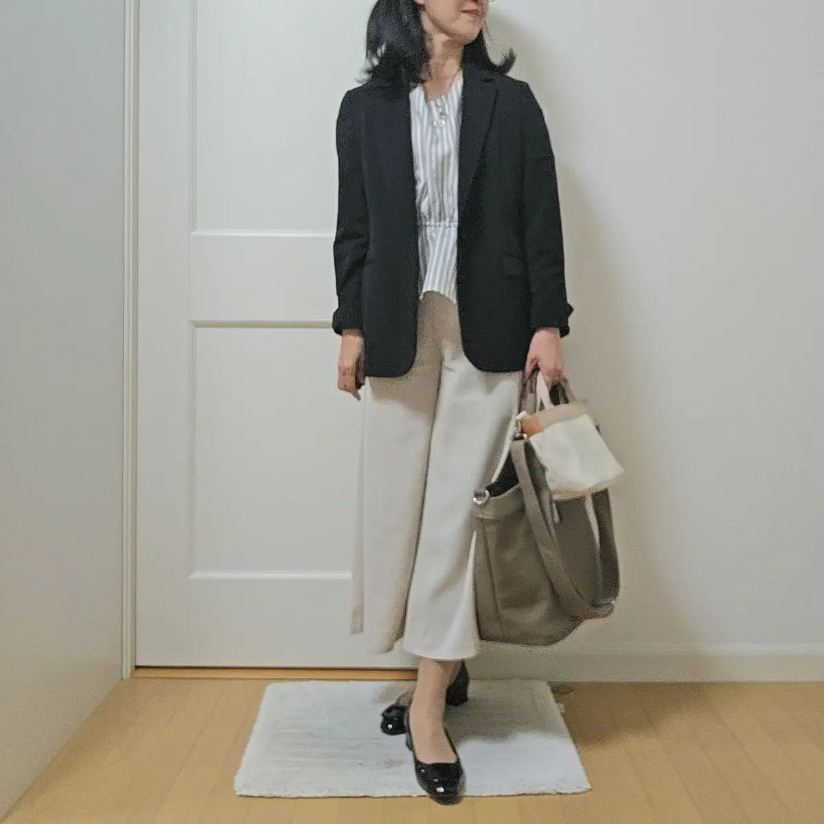 女性が黒のブレザージャケットとベージュのワイドパンツを着用し、グレーのトートバッグを手にしています。白い壁と木製の床が背景です。
