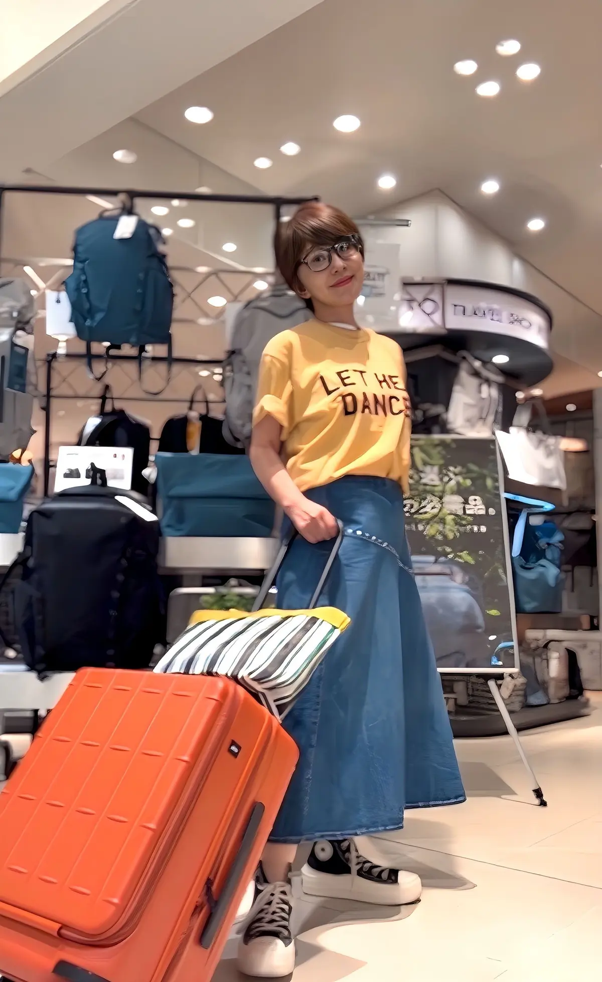 黄色いTシャツとデニースカートを着た女性が、空港や旅行用品店でオレンジ色のスーツケースとストライプ柄のトートバッグを手に持っています。背景にはさまざまなカバンや旅行用品が並んでいます。