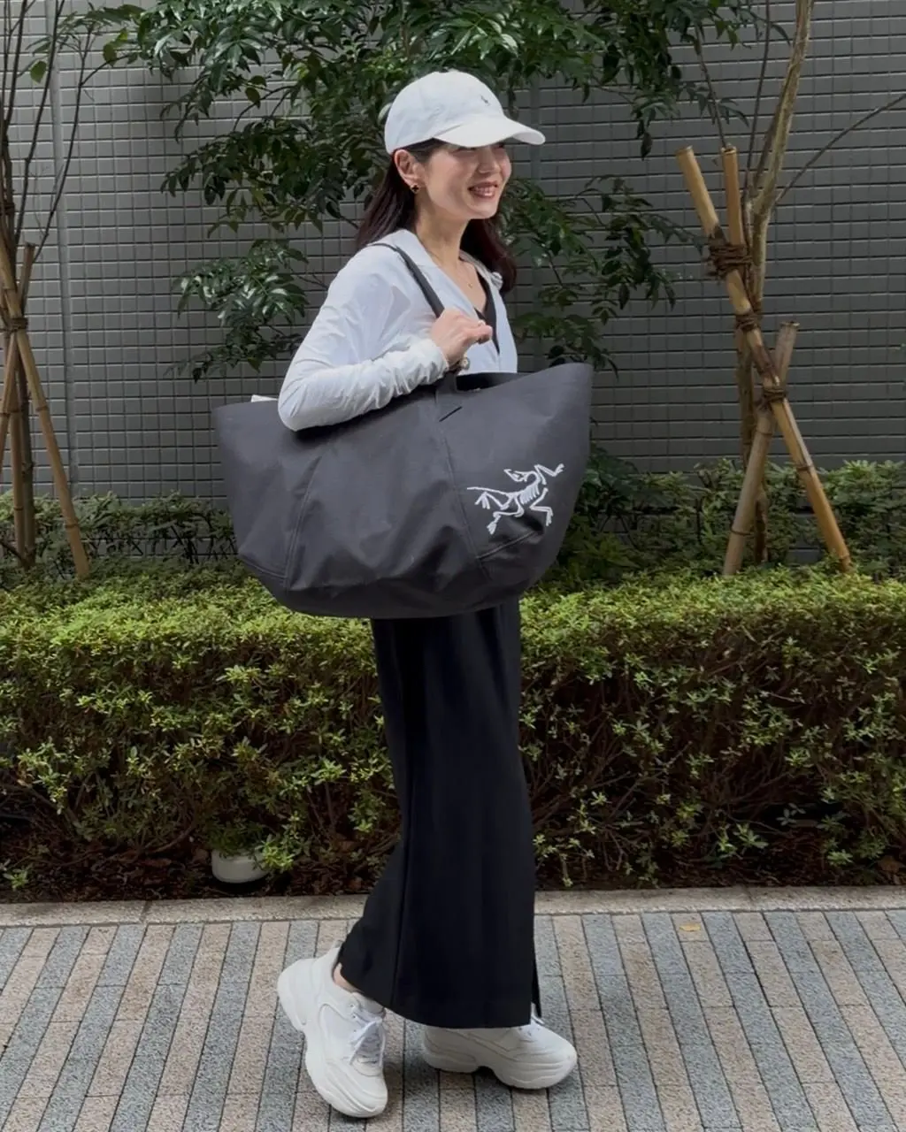 白いキャップと長袖シャツ、黒いロングスカートを着用した女性が、グレーのトートバッグを肩にかけ、笑顔で歩いている。背景には緑の植栽と灰色の壁がある。足元は白いスニーカー。