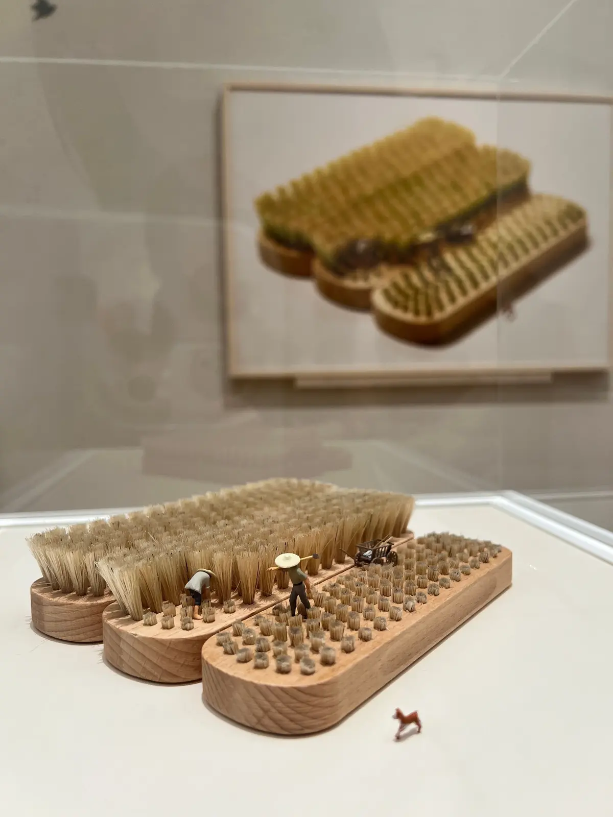 木製のブラシの上に、ミニチュアの農作業をする人々が配置されたアート作品。背景には、同じモチーフの額縁に入った写真が飾られている。