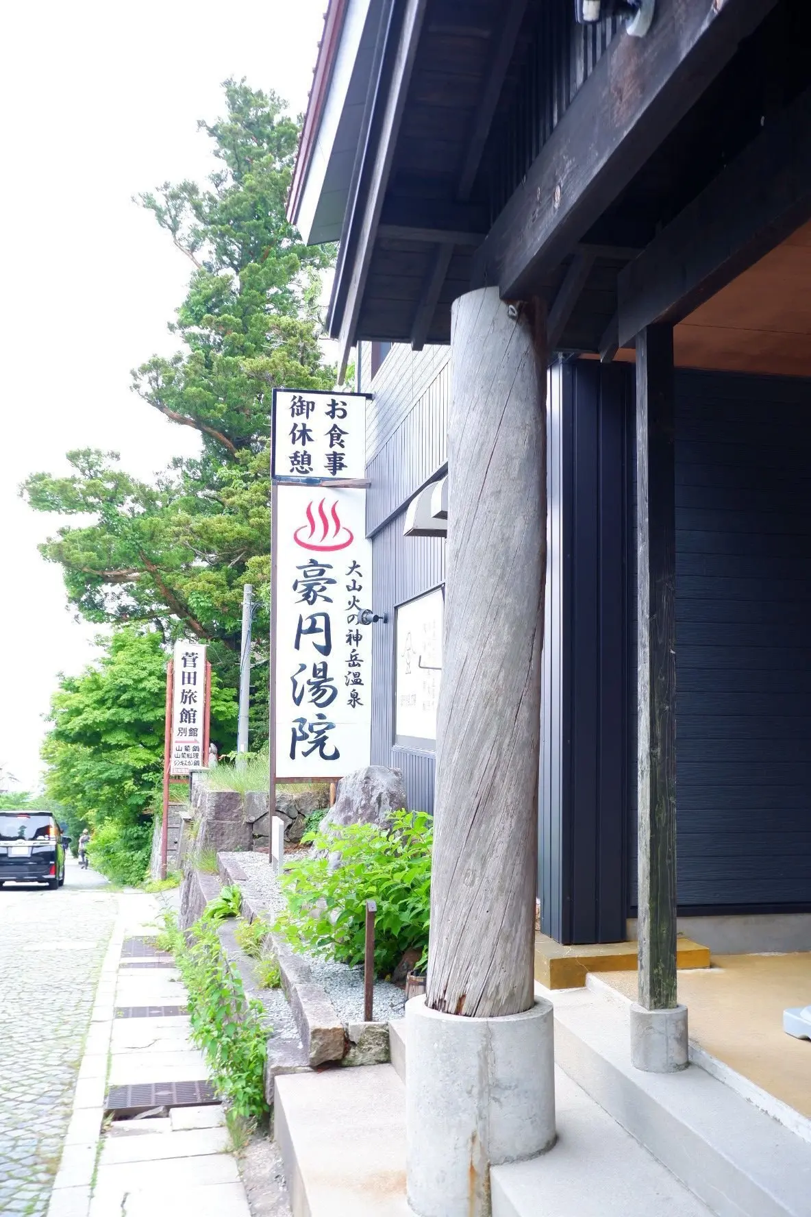 木造の建物の入り口に「豪円湯院」と書かれた看板があり、緑豊かな木々が背景に生えている