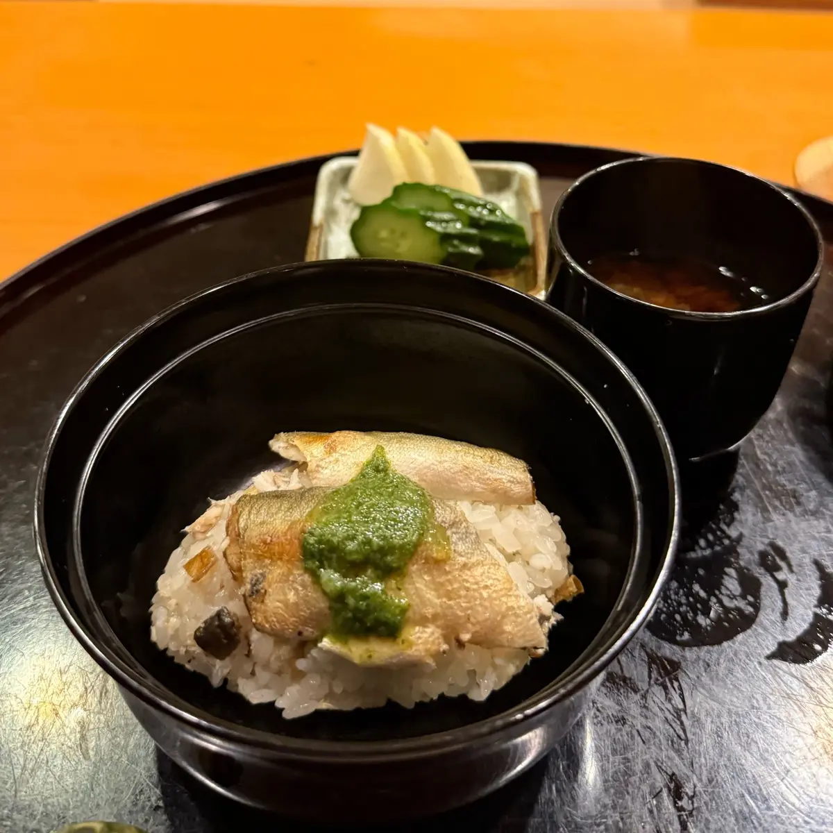 黒い漆塗りの漆器盆に盛り付けられた、サバの塩焼きとご飯の丼。ご飯の上には、刻みネギとワサビを混ぜた緑色の薬味が乗っている。盆には、白菜とキュウリの漬物、黒い汁椀も添えられている。背景はオレンジ色。
