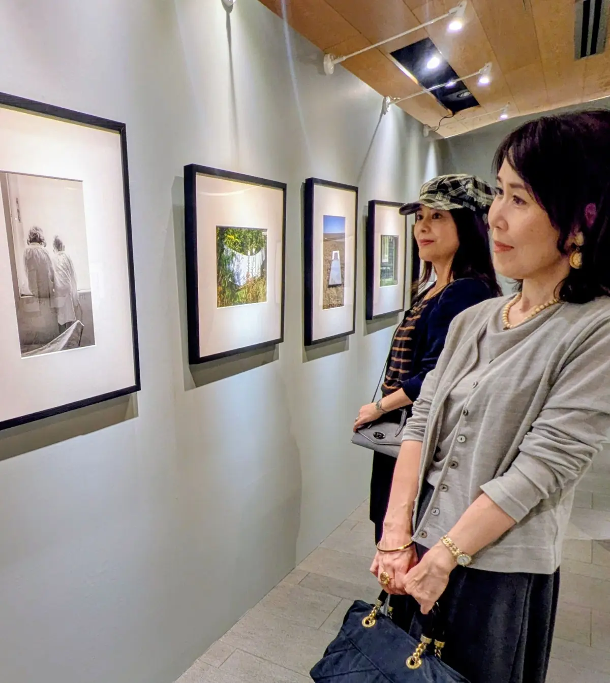 美術館で写真作品を鑑賞する2人の女性。左には3枚、右には2枚の額装された写真が飾られており、女性たちはそれらを眺めている。女性は黒いバッグを持ち、金色のアクセサリーを身に着けている。