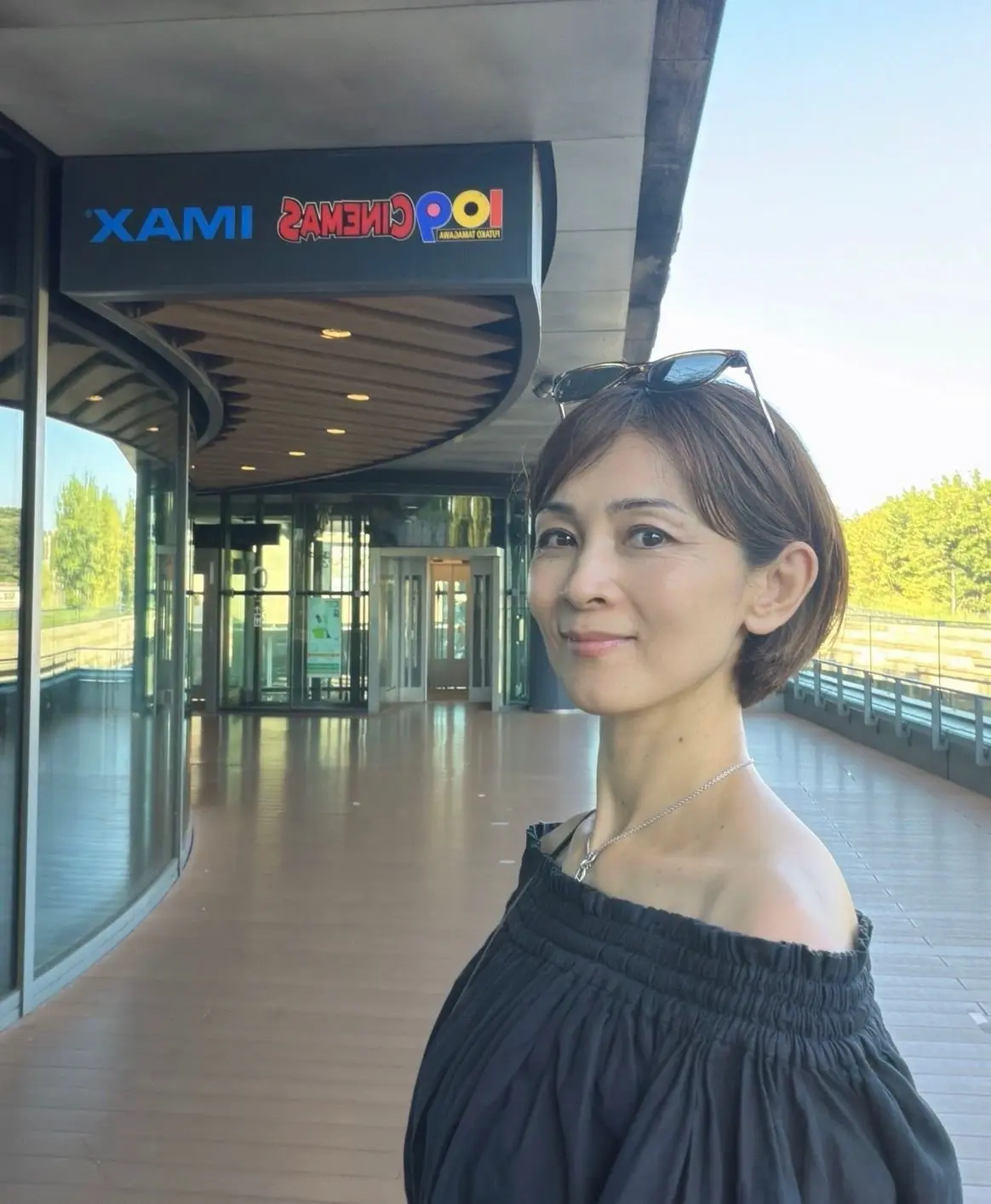 黒いトップスを着た女性が、XAMI 2AMEI POOLの入り口付近でこちらを見ている。背景にはガラス張りの通路と緑豊かな景色が広がる。女性はサングラスをかけ、右耳にはピアスをしている。