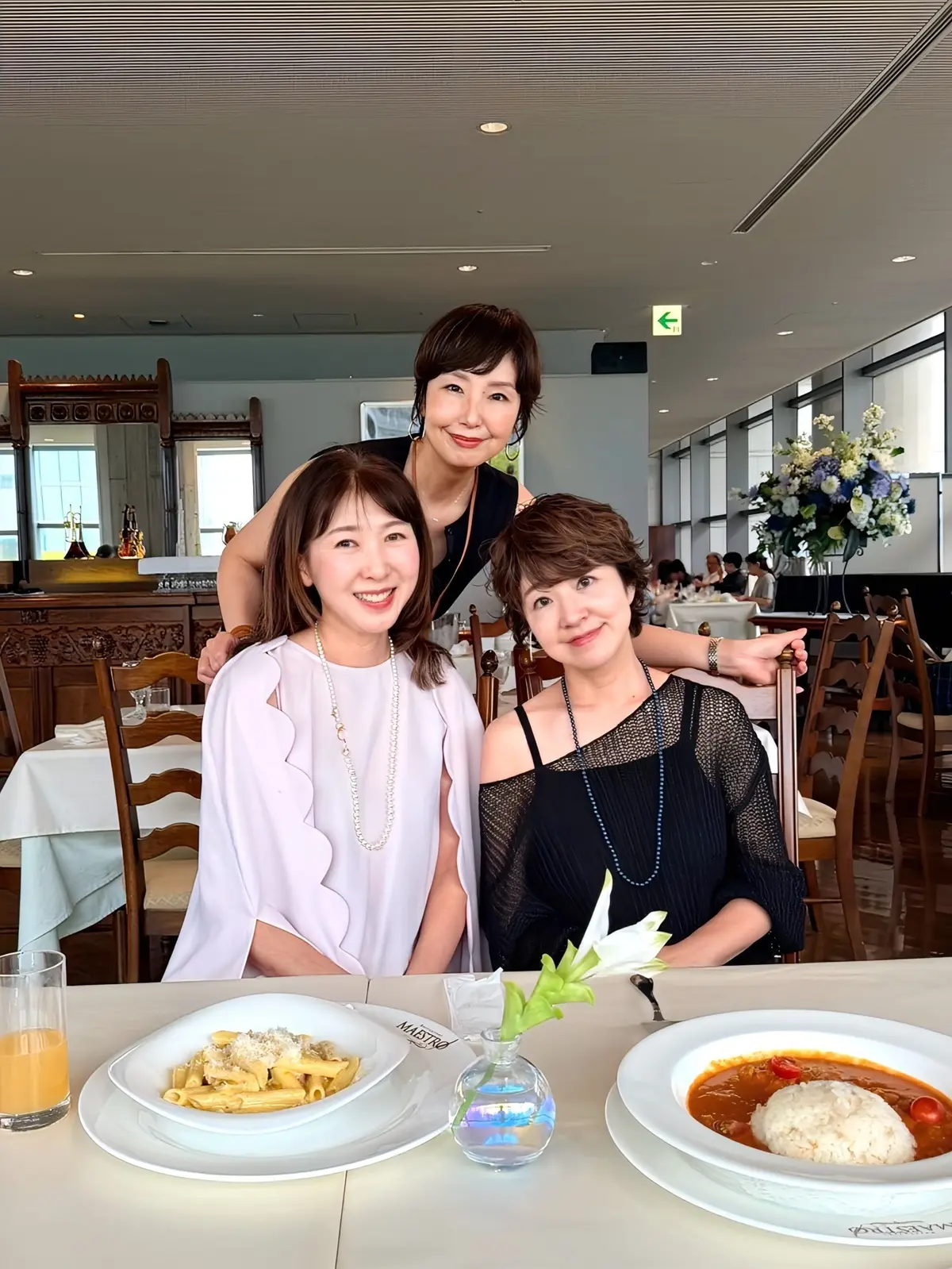 レストランで食事をする3人の女性。左は淡いピンクのワンピース、中央は黒のタンクトップ、右は黒の網目模様のトップスを着ている。それぞれ笑顔でカメラを見ている。テーブルにはパスタとカレーが盛り付けられている。背景には窓から光が差し込み、花が飾られている。