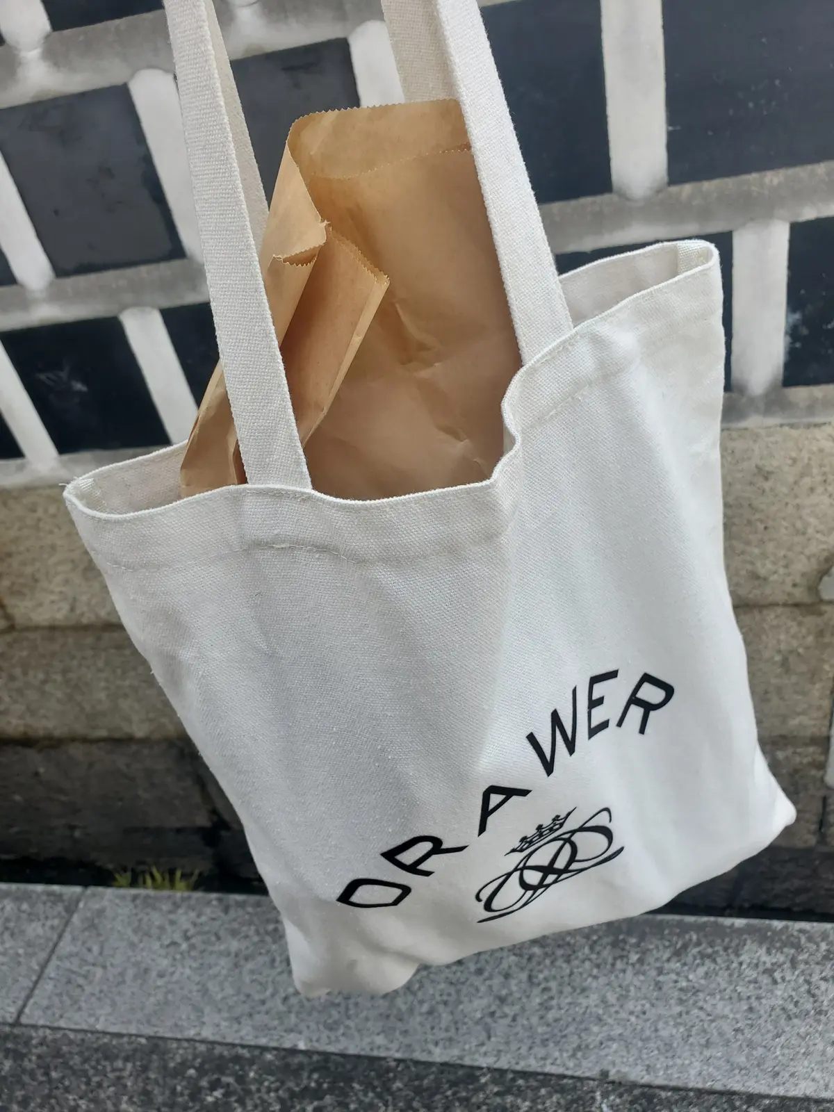 白地のトートバッグに、茶色の紙袋が一つ入っている。バッグ正面下部には、黒色の「ORAWER」の文字と、王冠をモチーフにしたロゴマークがプリントされている。背景には、黒と白の格子模様のフェンスが見える。