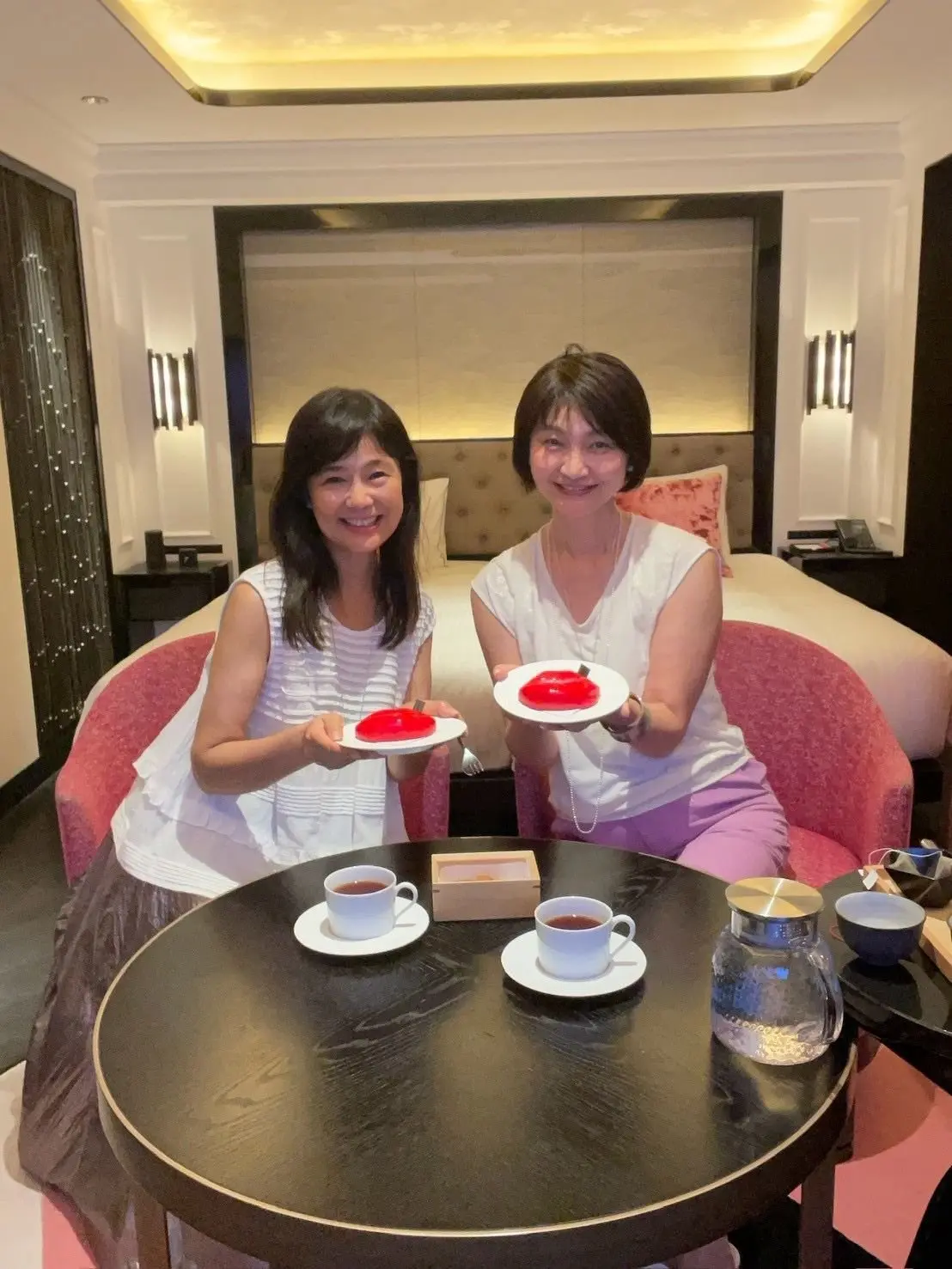 ホテルの部屋で、2人の女性が笑顔でデザート（赤いゼリー）とコーヒーを手にしている。丸いテーブルを挟んで座り、背景にはベッドと装飾が配置されている。