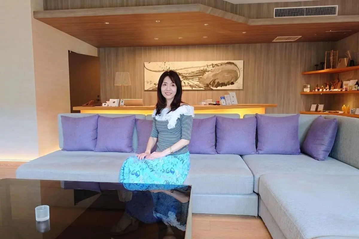 三輪湯河原のロビー。女性がグレーのソファに座っている。女性は青と白の柄のスカートと、白のレース襟のボーダーシャツを着ている。背景には、ベージュの壁掛けアートと、棚に並んだ商品が置かれている。