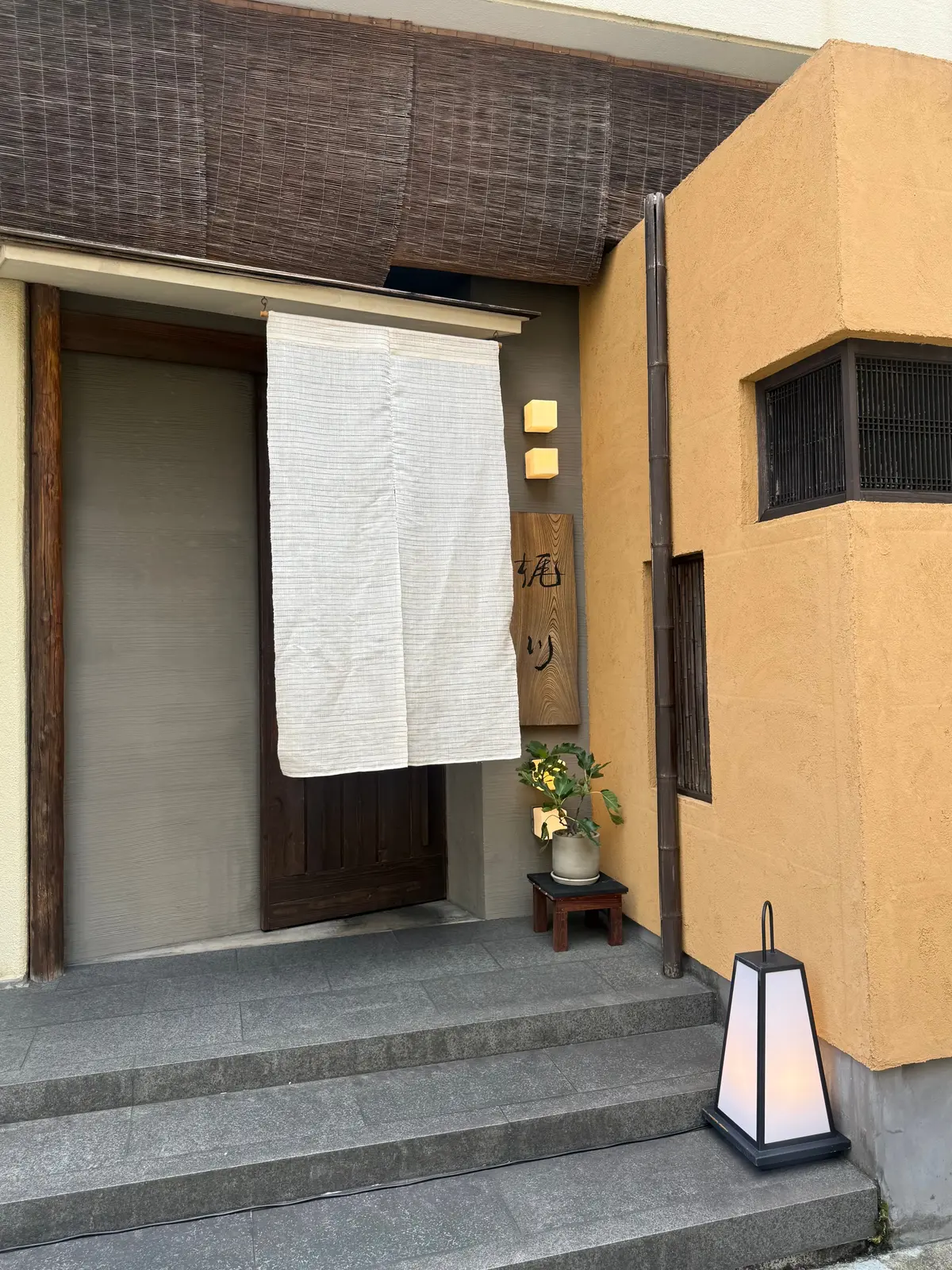 石造りの階段と、障子戸の入り口。白い布が掛けられ、右には木製の看板と植物が置かれている。壁は黄土色で、格子窓がある。足元には灯籠が置かれている。