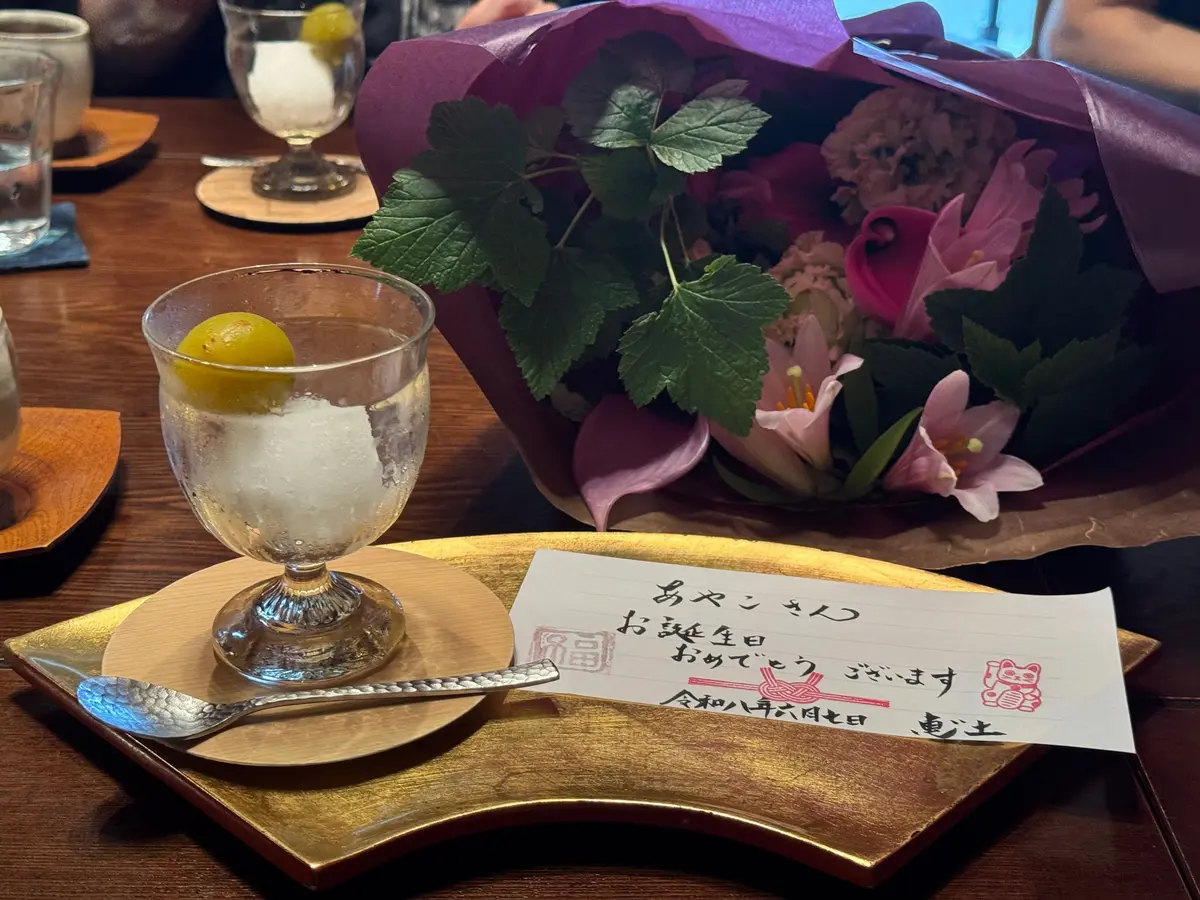 紫色のリボンが巻かれた花束と、氷とレモンの入ったガラスカップが木製のテーブルに置かれています。金色のプレートに日本語のカードとスプーンがあります。