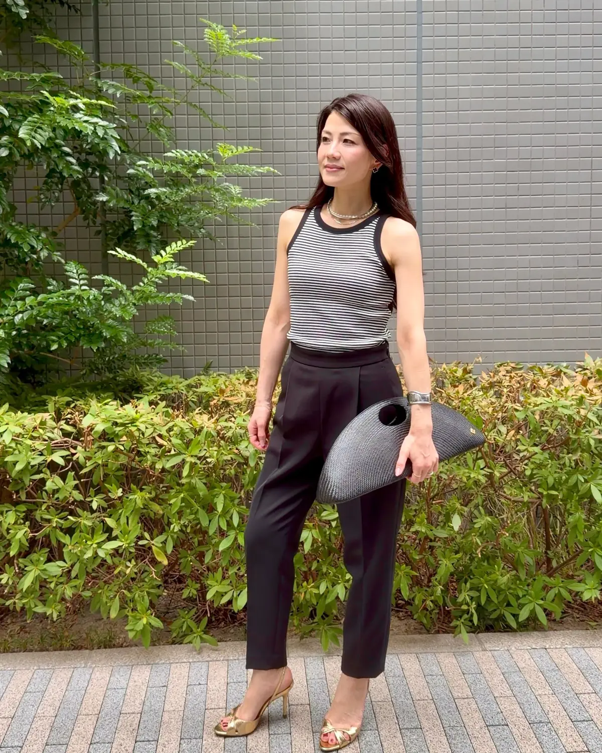 黒と白ボーダーのタンクトップに黒いパンツ、ゴールドのパンプスを着用した女性。右手に黒いクラッチバッグを持ち、背景には緑の植栽とグレーの壁がある。