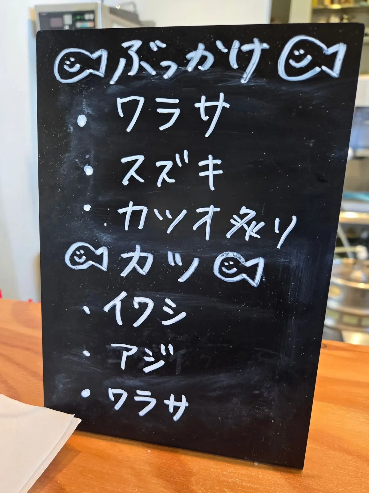 黒板に手書きで「バラカケ」と書かれたメニュー。その下に、クラサ、スズキ、カツオネリ、カツ、イワシ、アジ、クラサと列挙されている。各項目の前には笑顔の顔のマークが付いている。