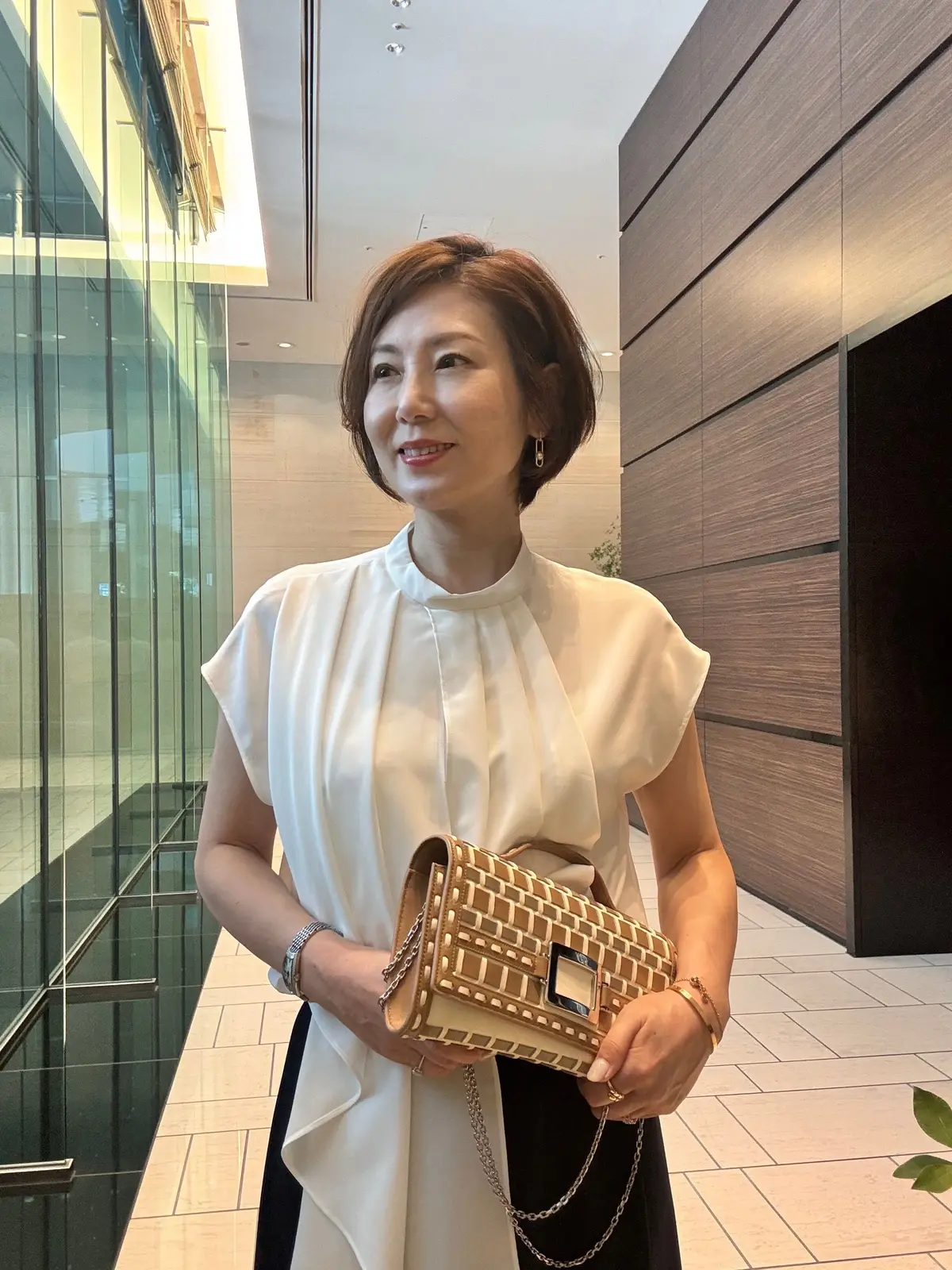白いトップスに黒いスカートを着用した女性が、エレベーターホールのタイルの床に立っている。右手に茶色のバッグを持ち、微笑んでいる。短い茶色の髪型で、耳には控えめなピアスをしている。
